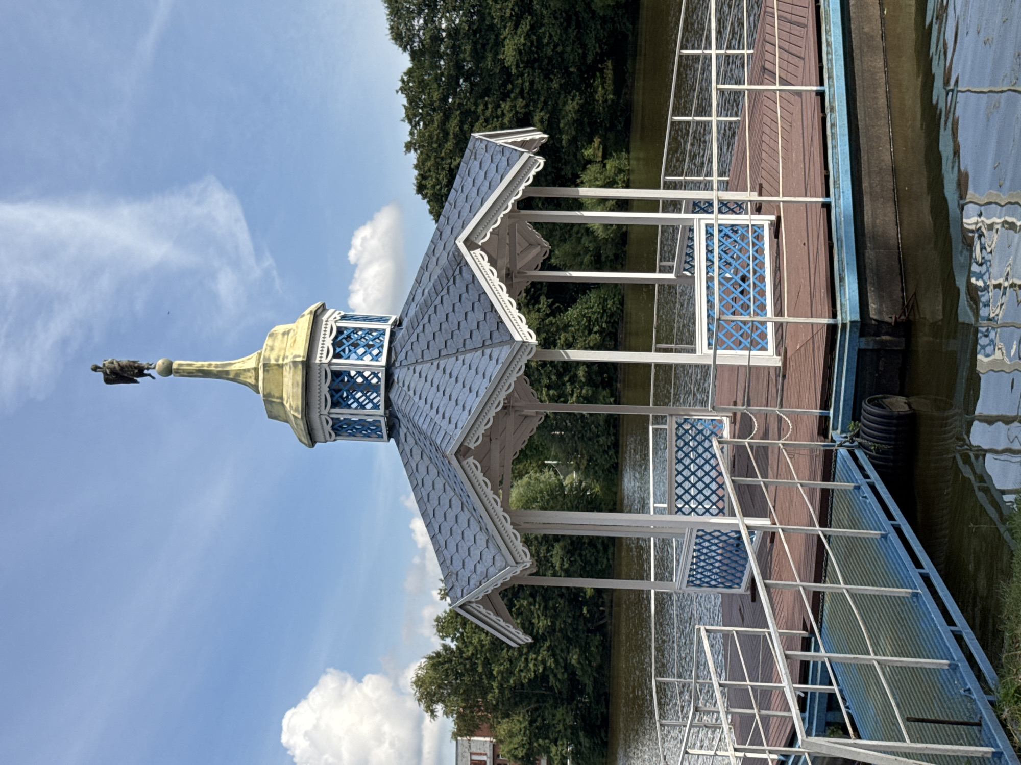 A picturesque wooden gazebo with a blue, shingled roof and white ornate trim stands on a floating dock. The gazebo is topped with a golden spire crowned by a black statue of a bird, possibly an eagle. The dock itself is made of brown wooden planks and has white metal railings. A blue ramp leads from the shore to the dock. The scene is set against a backdrop of a calm, dark body of water and lush green trees. The sky is blue with scattered white clouds. The gazebo is located in Sestroretsk, Russia, likely in one of its parks or recreational areas.