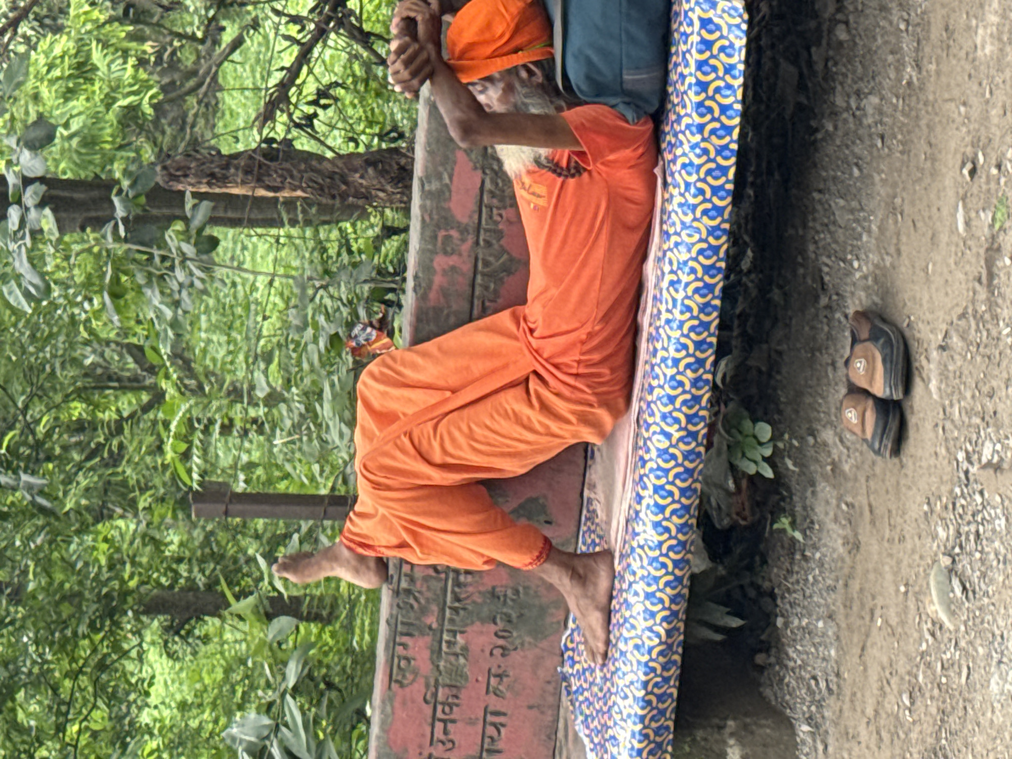 An elderly man wearing an orange robe is lying on his back on a bench in Rishikesh, India. His legs are extended and his right foot is resting on the back of the bench. His hands are clasped behind his head, and his eyes are closed. A blue pillow is under his head. The bench is covered with a blue and yellow patterned mat. There are trees and foliage in the background, suggesting an outdoor setting. A pair of sandals are placed on the ground in front of the bench. The bench has inscriptions in a language that appears to be Hindi.