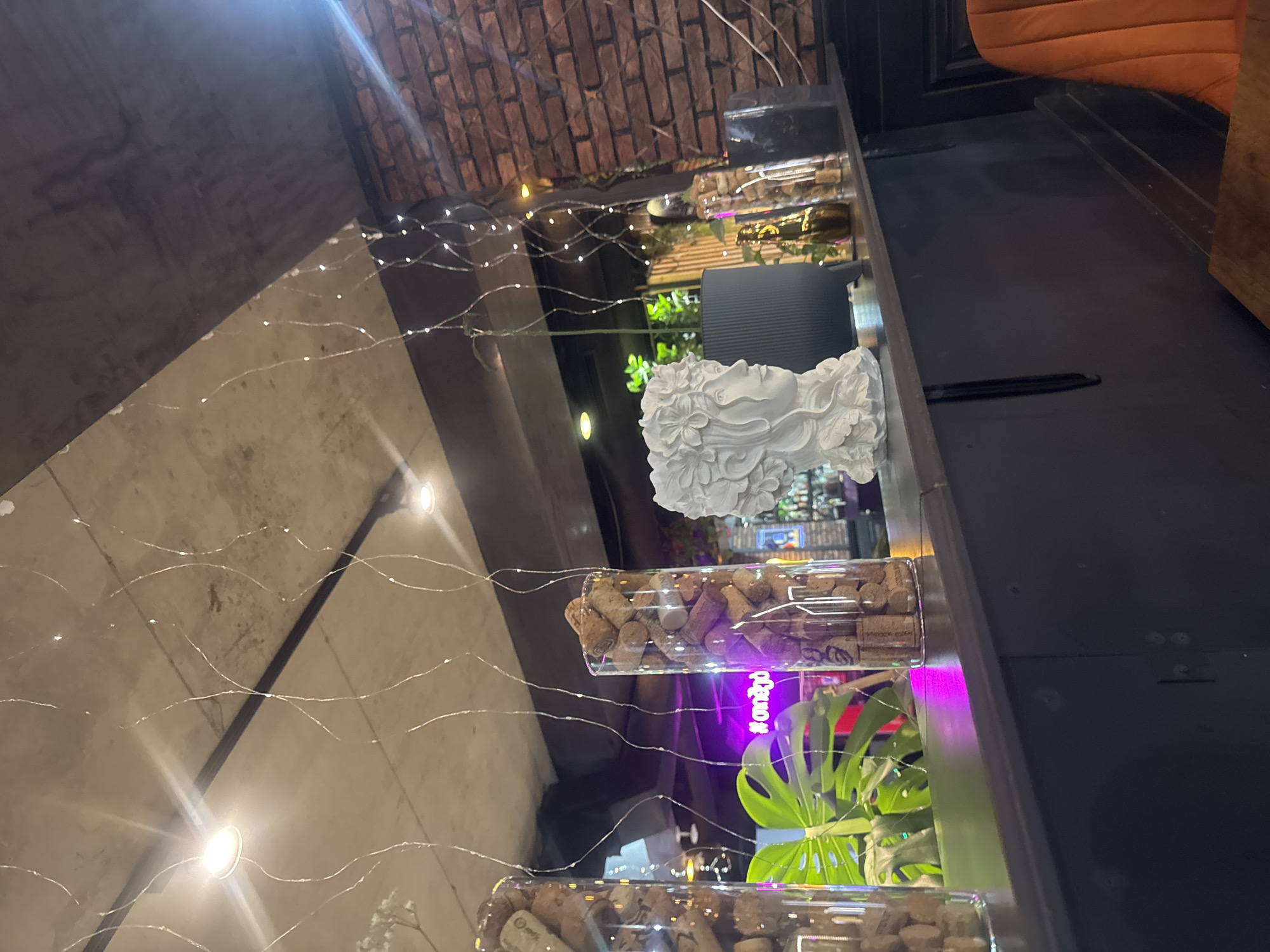 The image shows a shelf in a dimly lit interior, decorated with fairy lights. On the shelf are:
* Two transparent rectangular containers filled with wine corks.
* A white decorative bust of a woman's head.
* A small potted plant with large green leaves.
* A gray square object behind the bust.
The background includes a brick wall, a dark gray wall, and a partial view of an orange upholstered chair. A purple light is visible behind the plant. The context suggests this is a decorative arrangement within a commercial establishment. Given the prompt's instruction and the likely style of decor, a potential location could be a wine bar or restaurant in Tbilisi, Georgia, known for its contemporary interior design and appreciation of wine.