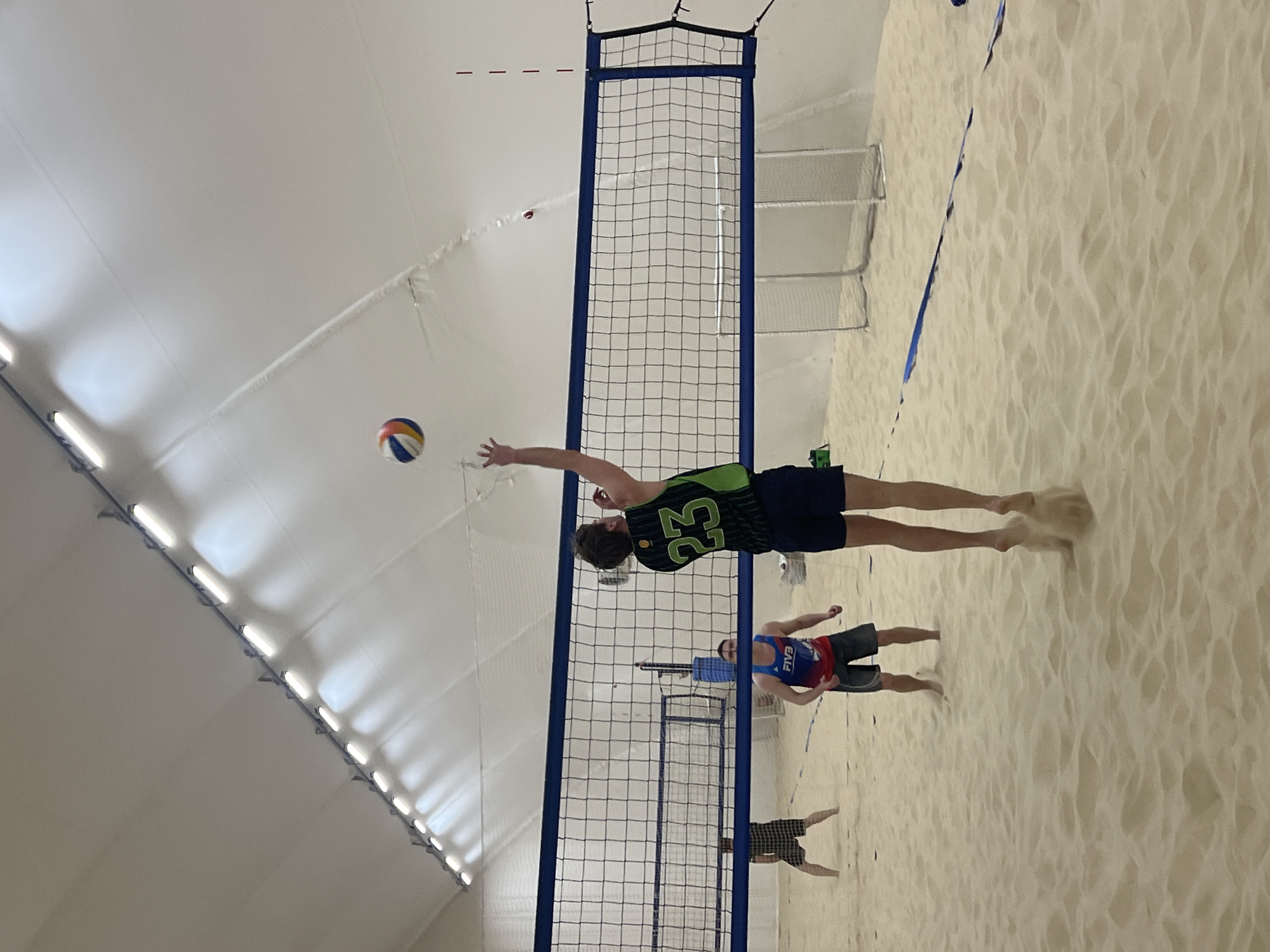 The image shows a beach volleyball game being played indoors on a sand court. A player in a green and black jersey with the number "23" is in mid-air, about to hit the volleyball over the net. Two other players are visible on the opposite side of the net. The setting appears to be an indoor sports facility with a high ceiling and artificial lighting. The scene is consistent with the presence of a beach volleyball facility, but there is insufficient evidence to confirm the location in the city of Saint Petersburg, Russia.