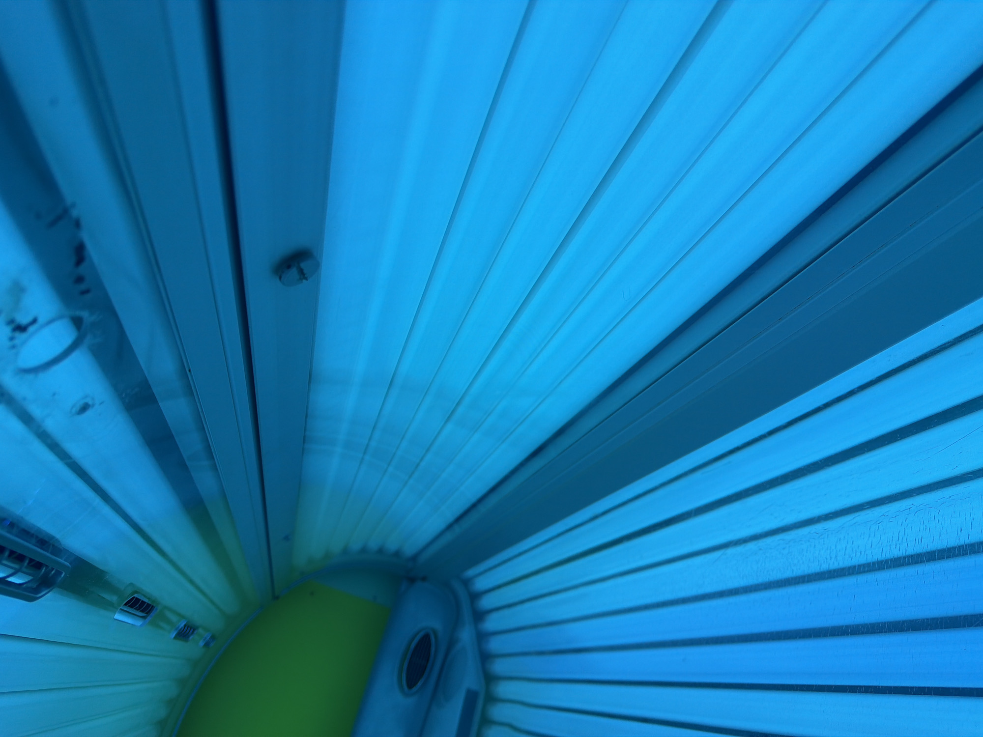The image shows the interior of a tanning bed. The bed is illuminated by bright blue UV lights, which are arranged in parallel lines along the ceiling and the sides of the bed. The interior of the bed is curved, and the lighting creates a sense of depth and immersion. There are no people visible in the image. The overall impression is one of a sterile, artificial environment. The location in Kyiv, Ukraine is not discernible from the image itself.