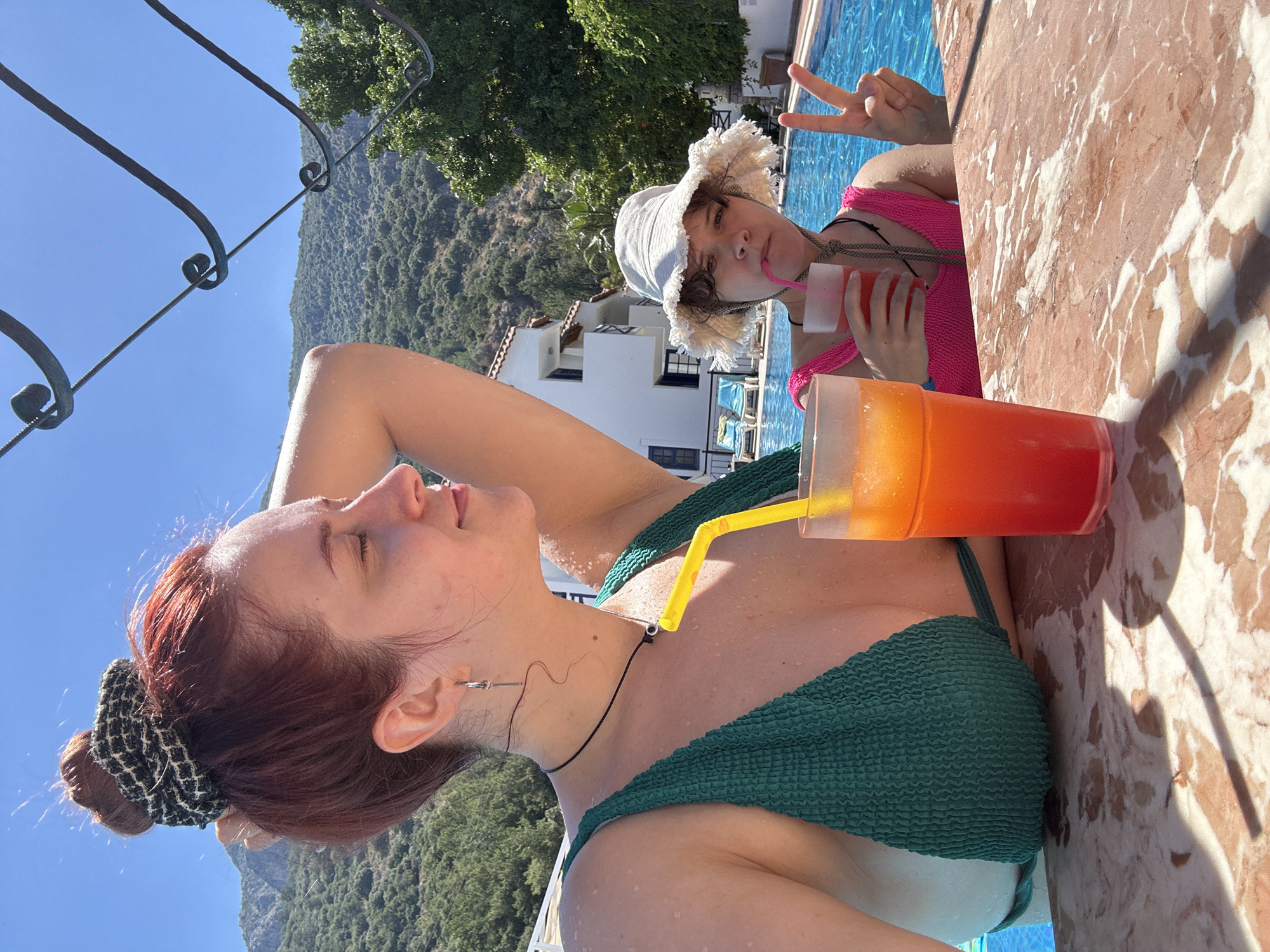 This media file depicts two individuals relaxing by a swimming pool on a sunny day, likely in Fethiye, Turkey, given the mountainous and coastal resort context. The image is taken from a low angle and rotated 90 degrees clockwise.
In the foreground, a young woman with reddish-brown hair, tied back with a black and white patterned scrunchie, is seen in profile. She wears a dark green, textured bikini top and has water droplets on her skin, suggesting she has been in the pool. She holds a large clear cup with a layered orange and red drink, complete with a yellow straw. Her eyes are closed, or looking downwards, indicating a relaxed state.
In the background, partially obscured by the foreground subject and a marble-patterned poolside surface, another person, possibly younger, is visible in the swimming pool. They wear a bright pink swimsuit and a white fringe-style bucket hat. This person looks towards the camera, sipping from a clear cup with a pink drink and a pink straw, and makes a peace sign with their left hand, suggesting a playful and happy mood.
The setting features clear blue skies, lush green, tree-covered mountains in the distance, and white buildings with red-tiled roofs, characteristic of Mediterranean architecture. Metal railings with ornate curls are also visible, possibly part of a balcony or pool barrier. The bright sunlight suggests it is daytime. There is no visible text in the image. The overall impression is one of a relaxing vacation by the pool in a scenic location.