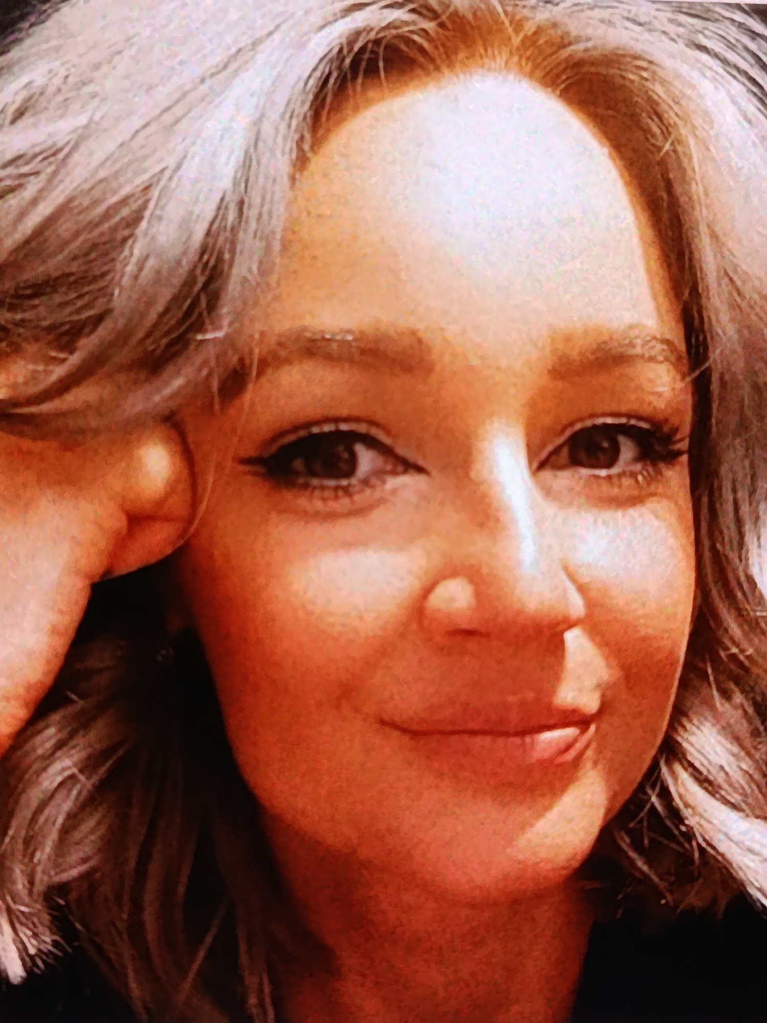 The image is a close-up portrait of a woman with blonde, wavy hair. She is looking towards the viewer with a slight smile. Her right hand is resting on her cheek, and her left arm is visible at the bottom of the frame. The woman is wearing makeup, including eyeliner and mascara. The lighting suggests an indoor setting, possibly a home or a studio. The background is dark and indistinct. No specific location in Bălți, Moldova, can be identified from this image.