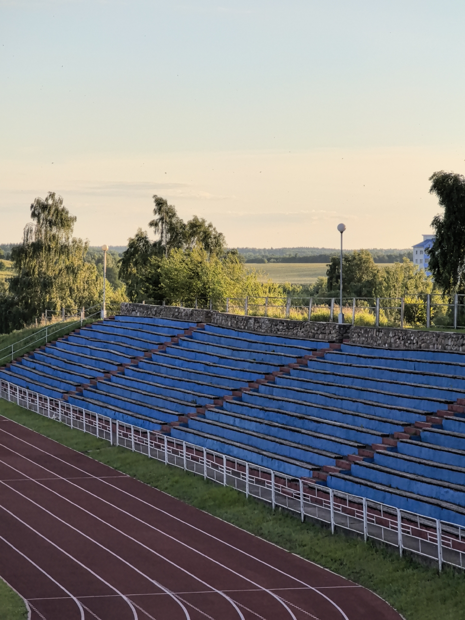The image depicts an outdoor stadium with blue bleacher seating, a running track, and a distant landscape under a clear sky. The bleachers are empty and arranged in rows, with a stone wall at the base of the stands. A white metal railing separates the track from the seating area. The running track itself is a dark reddish-brown color with white lane markings.
In the background, there are trees, fields, and a blue building. A single lamppost stands near the bleachers. The sky is a pale blue with hints of yellow, suggesting it is either early morning or late afternoon. The overall scene is serene and empty, with no people or activities visible. The lighting suggests a calm and quiet atmosphere. The location is not identifiable from the image. There is no visible text.