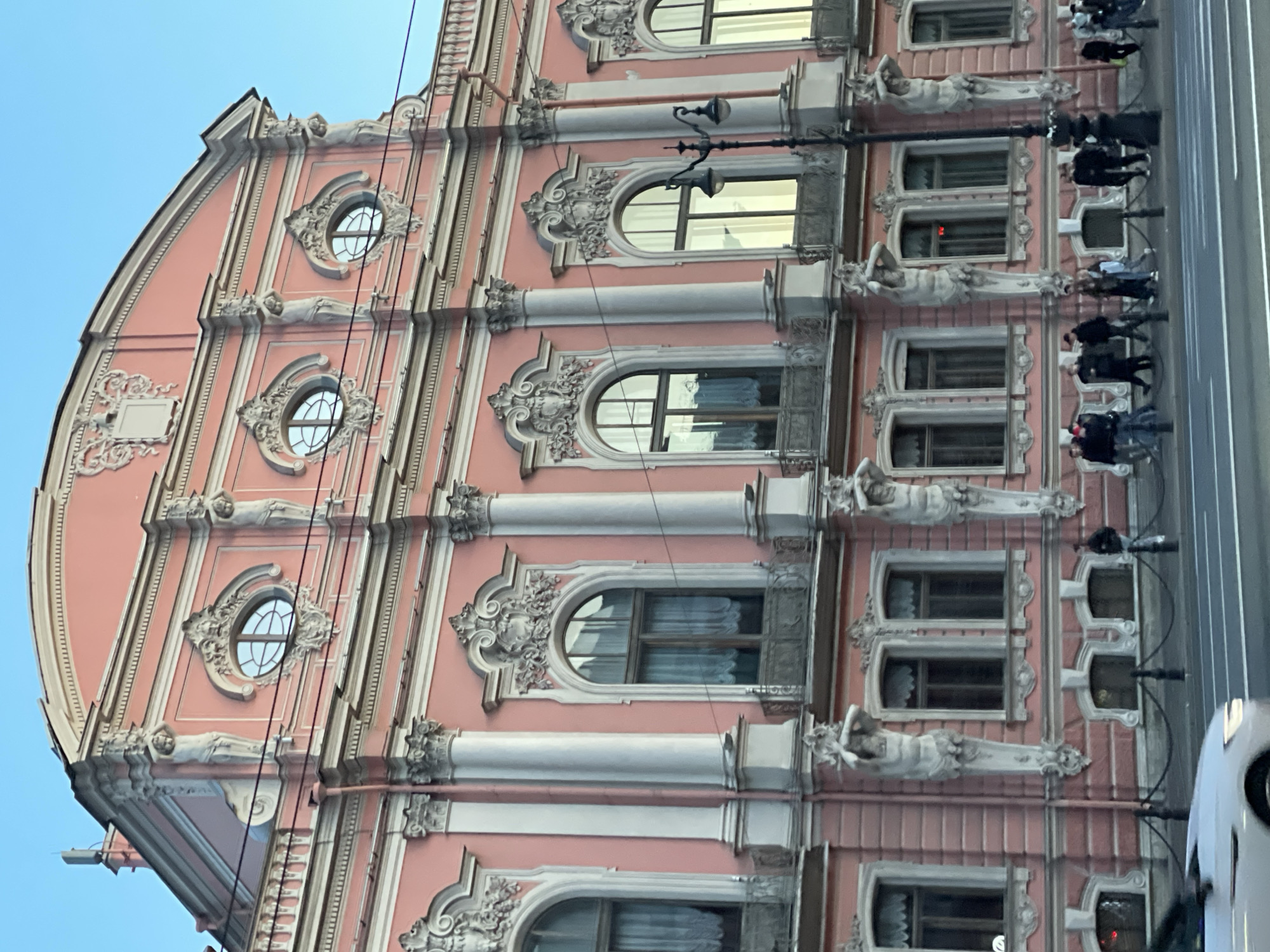 The image displays a close-up view of a highly ornate, multi-story building facade, rendered in a striking salmon pink color with extensive white decorative architectural elements. The perspective is from street level, looking upwards, and the image is rotated 90 degrees clockwise from its original capture, making the building appear to lean left.
The building exhibits a rich architectural style, possibly Art Nouveau or Baroque Revival, characterized by elaborate stucco ornamentation. Prominent features include:
- Multiple rows of arched windows, rectangular windows, and smaller, ornate oval or round windows, each framed by intricate moldings, volutes, and decorative keystones.
- White pilasters and columns with decorative capitals that punctuate the facade vertically.
- Sculptural elements, specifically figures resembling caryatids or atlantes, depicted as reclining or draped figures, supporting various sections of the facade between windows.
- Balustrades and decorative railings on some window sills.
- A distinctive curved gable or roofline at the top, also adorned with white moldings.
- The top section of the building features decorative plaques and additional sculpted figures.
In the foreground (right side of the rotated image), a busy street scene is partially visible. A gray road with painted lines suggests a multi-lane thoroughfare. A white car is partially seen on the right, and several pedestrians are walking on a sidewalk alongside the road. Black streetlights with decorative lamps are present, and utility lines or cables crisscross in front of the building.
The sky in the upper left corner (top in the original orientation) is clear and blue, indicating daytime. No legible text is visible in the image. The architectural style and busy urban setting are consistent with Saint Petersburg, Russia.