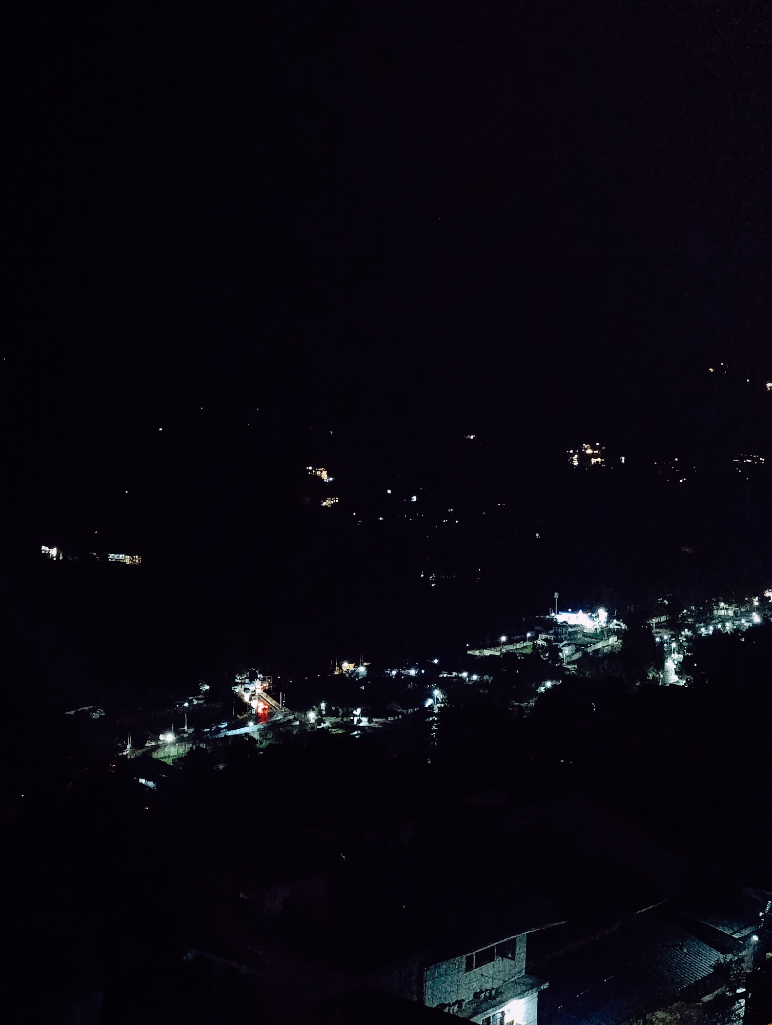 The image is a nighttime aerial view of Vashisht, India, showing a village nestled in a valley illuminated by numerous lights. The scene is predominantly dark, with the village lights creating a stark contrast against the blackness. In the foreground, the rooftops of several buildings are visible, some with illuminated windows. Further into the scene, a road winds through the village, lined with streetlights and buildings emitting a warm glow. Cars are visible on the road, their headlights and taillights creating streaks of light. In the background, more lights indicate the extent of the village and potentially other settlements in the surrounding hills. The overall impression is one of a vibrant community existing within a dark, mountainous landscape.