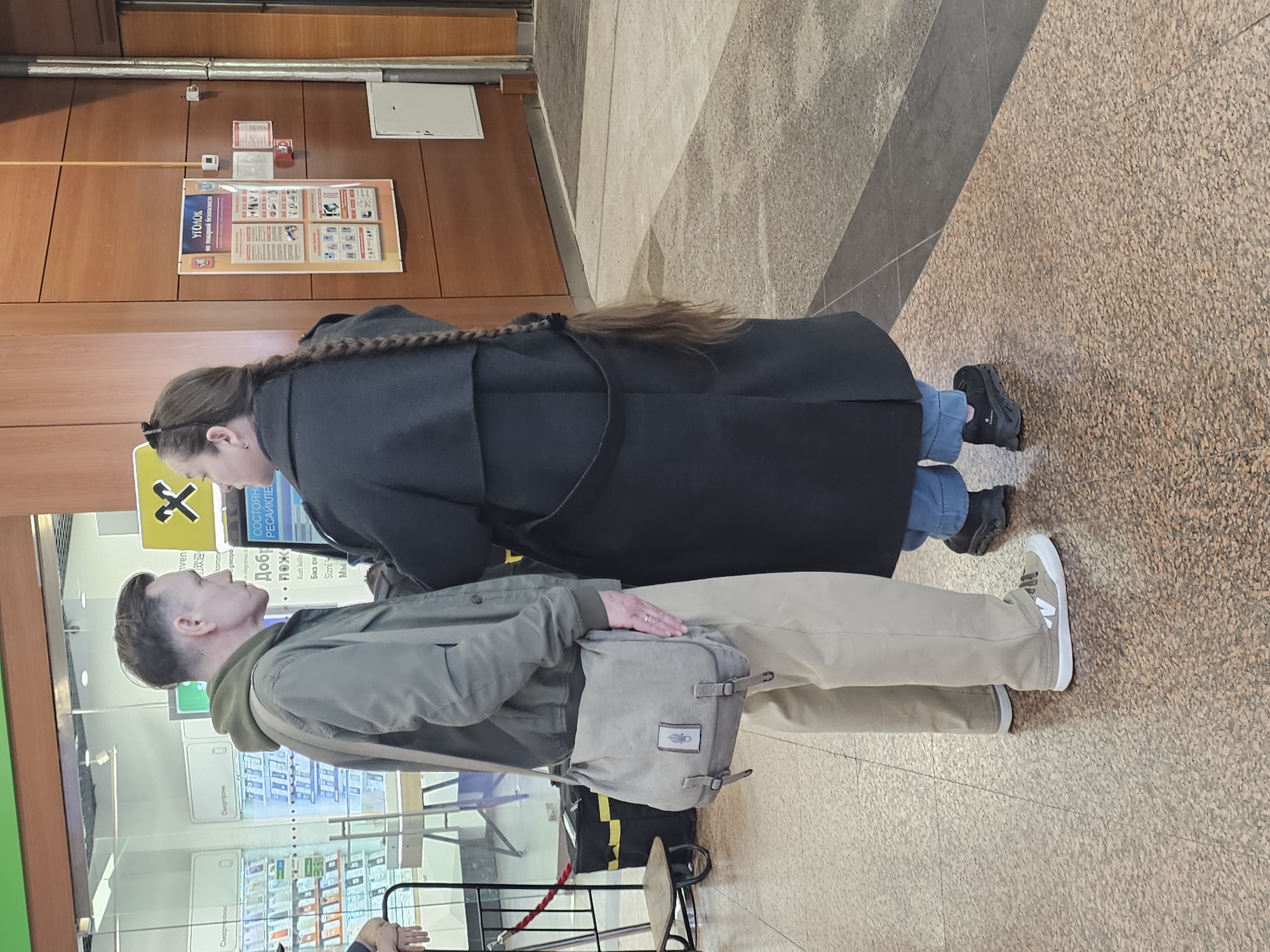 Here is a detailed description of the image:
The image appears to be taken indoors, possibly inside a transportation hub like an airport or train station in an area where Russian is spoken, based on the signage.
Two people are in the foreground. One is a woman wearing a long black coat with a braid in her hair. She appears to be looking at something to her left. Next to her is a man with short hair, wearing a greenish jacket, khaki pants, and sneakers. He has a messenger bag slung over his shoulder, and he also appears to be looking at something to his left. Their posture suggests they might be together and engaged in a shared activity, such as reading a schedule or looking at a store.
Behind them is a glass wall, displaying various advertisements and information, including signs in Russian about fire safety and recycling ("ΥΓΟΛΟΚ по пожарной безопаснос" and "СМАртфоны"), indicating the possible location within Russia or a Russian-speaking region. There is also some text in another language, likely Turkish ("Xush kellier," "Биз си," "Sizni," "Мы").
The background shows a wooden wall with various signs and notices, and the floor is tiled with a patterned design. The overall lighting in the area seems adequate.