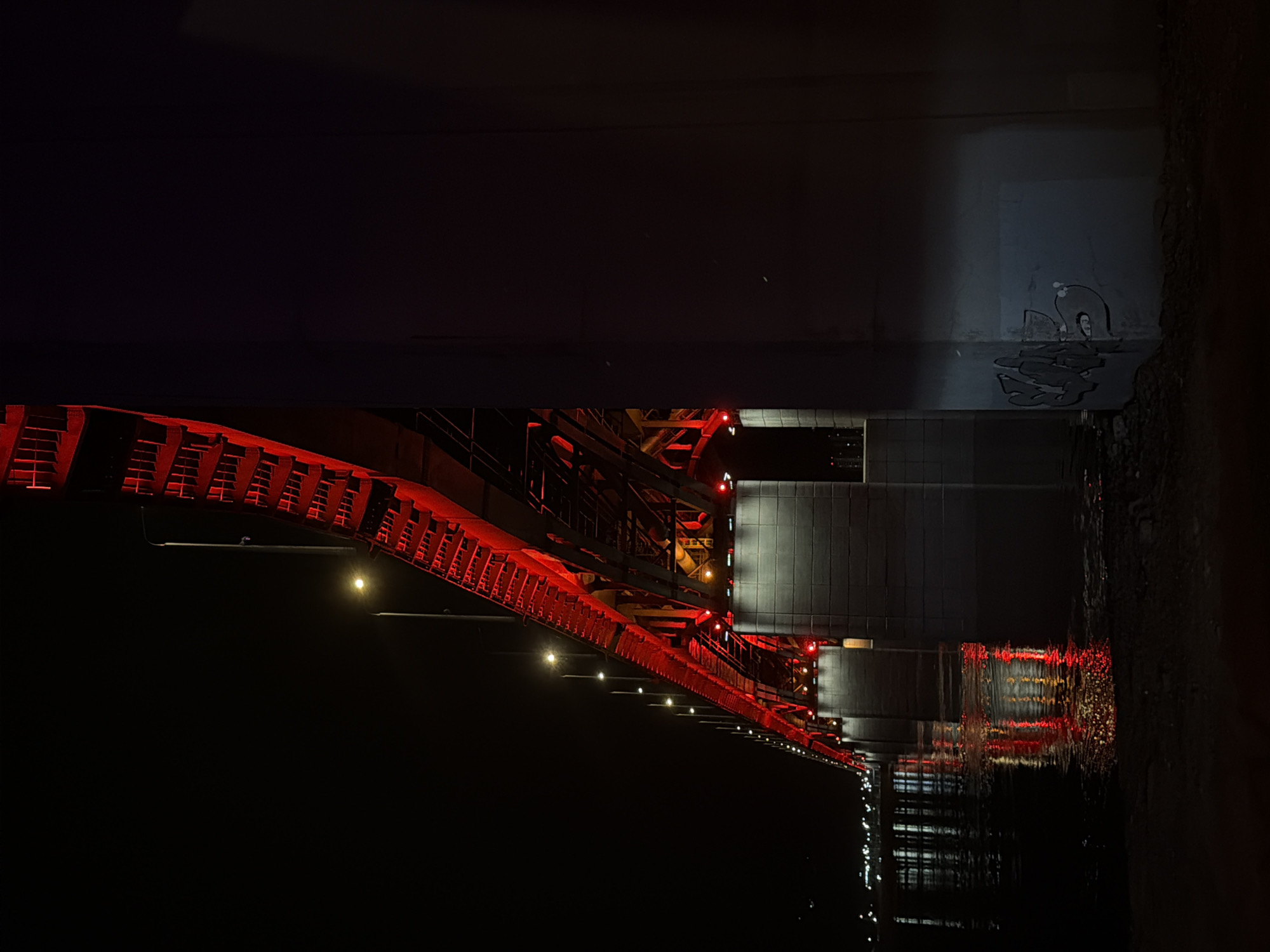 The image depicts a section of the Vinogradovsky Bridge in Krasnoyarsk, Russia, illuminated at night. The bridge is constructed of dark steel with red accents. The supporting structure is visible underneath the main span, with small lights running along its length. The bridge is reflected in the dark water below, which shows the reflections of the red lights. Graffiti is present on the bridge's support.