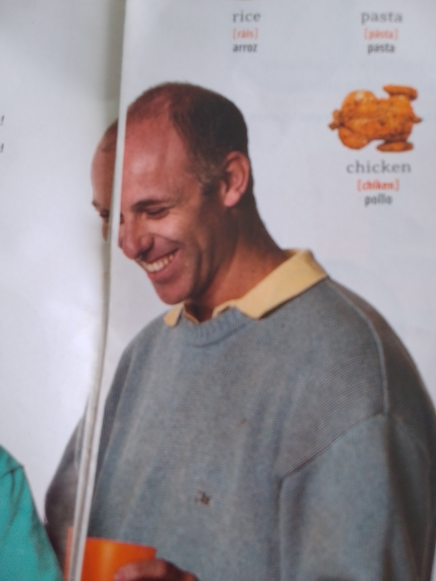 The image is a medium close-up shot of a middle-aged man with a shaved head, smiling broadly. He is wearing a grey crew-neck sweater over a yellow collared shirt. His head is tilted slightly, and his eyes are closed. In the background, on a white page, there is text related to food items. Specifically, the words "rice," "pasta," and "chicken" are visible, along with their phonetic pronunciations and translations in Spanish ("arroz," "pollo"). A small, golden-brown image of a roasted chicken is also present next to the word "chicken." To the left of the man, another person is partially visible, wearing a bright turquoise shirt, and holding an orange cup. The man's expression suggests a moment of amusement or happiness. The setting appears to be indoors, possibly a classroom or a home, given the educational content on the page and the presence of multiple people. There are no clear indicators of time of day or weather.