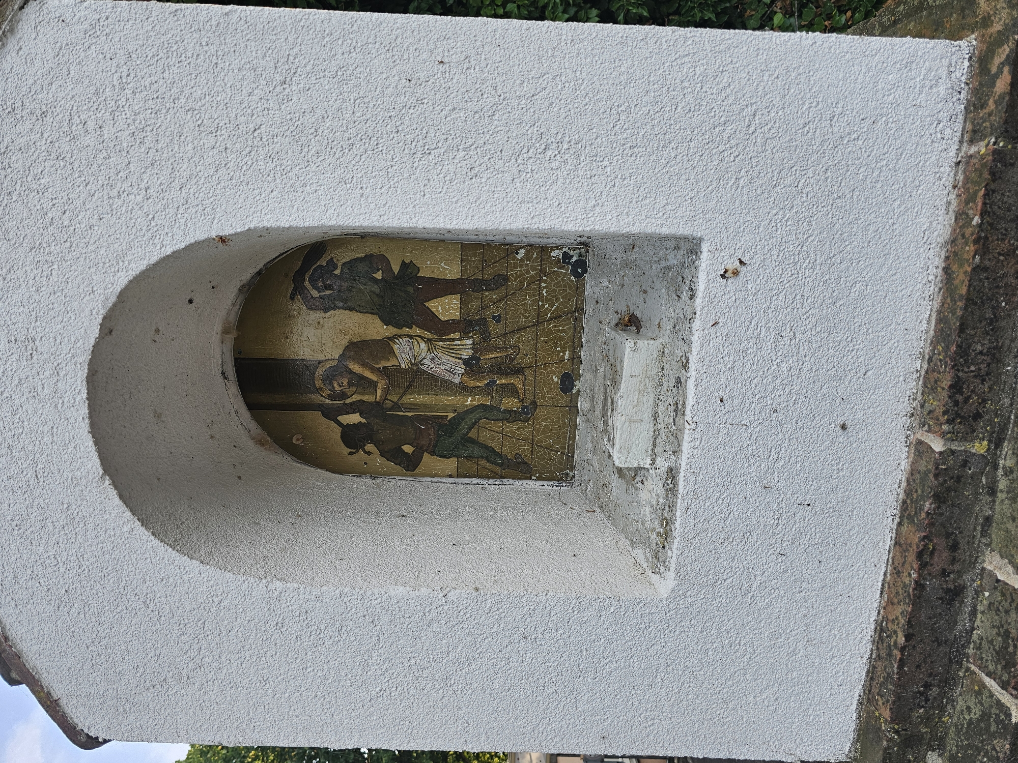 A small, arched niche in a white stucco structure contains a painting of three figures. The central figure is a man with a loincloth, who appears to be Jesus, bound to a vertical post. Two other figures, depicted with darker skin tones, stand on either side of him, wielding clubs or whips and striking him. The painting is aged and shows signs of cracking and wear. The niche is set into a weathered, white structure, possibly a wayside shrine, with a darker, brick-like base below. The background shows hints of greenery and a cloudy sky.