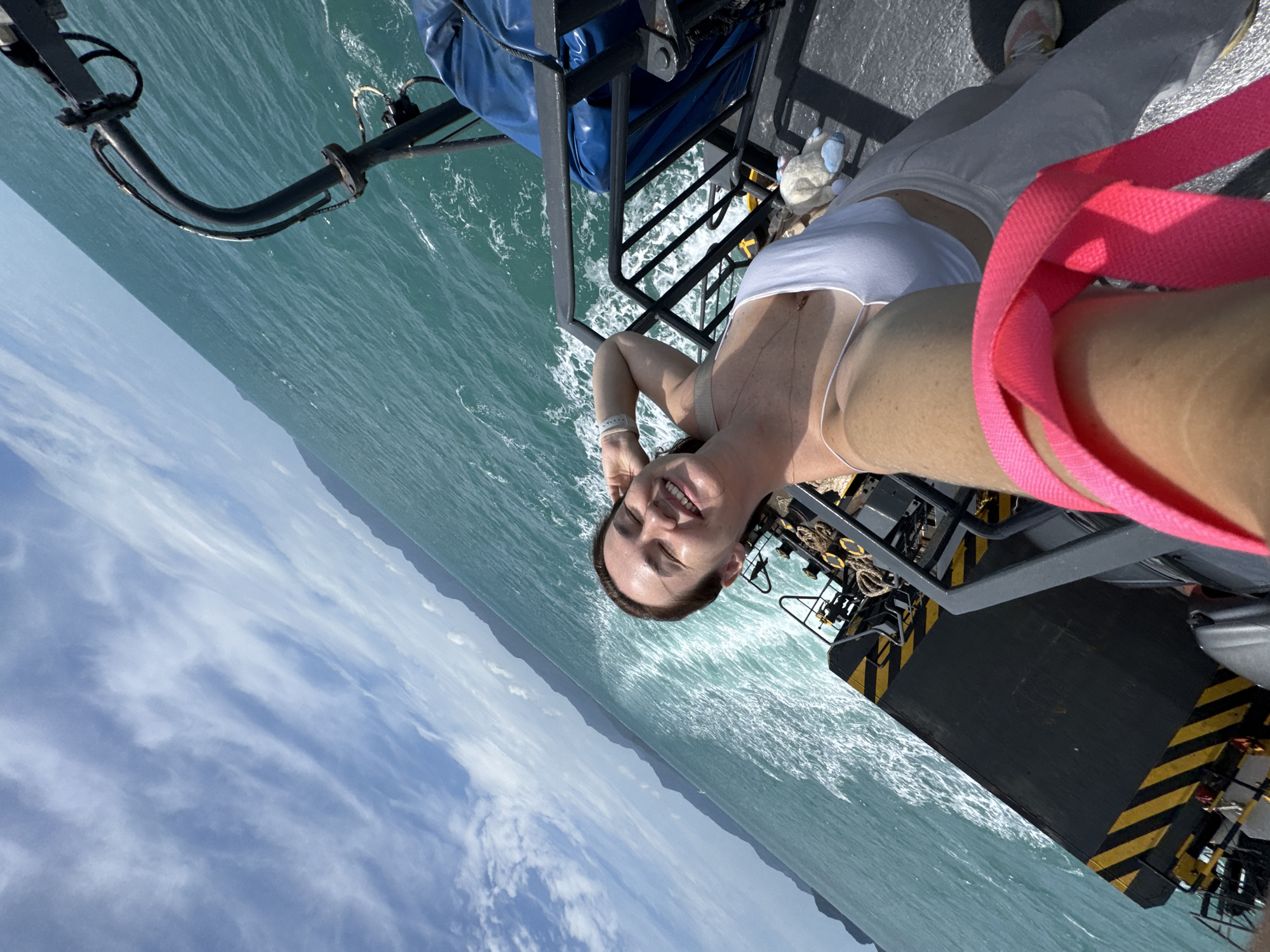 A woman with brown hair smiles as she takes a selfie on a ferry traveling on the ocean, possibly near Ko Pha-ngan, Thailand. She is wearing a white tank top and light-colored pants. Behind her, the turquoise ocean stretches to the horizon, with a distant coastline visible under a cloudy blue sky. The ferry deck is visible, with black and yellow striped markings and some equipment. A bright pink strap from a bag is in the foreground.