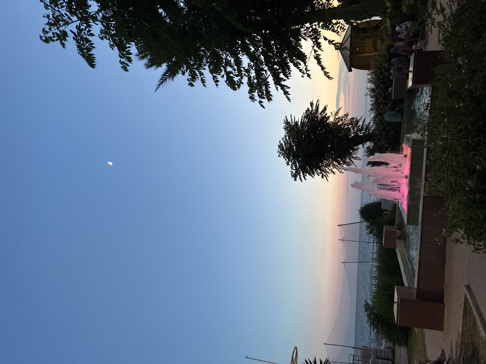 The image depicts a twilight scene at a resort or public area near the sea in Alanya, Turkey. A fountain with pink lighting is the central focus, with water jets spraying upwards. In the background, the calm sea stretches to the horizon, with a hazy sunset casting soft hues of pink and orange across the sky. Several trees, silhouetted against the fading light, frame the scene on the right and left. A small, yellow gazebo or shelter is visible on the right. Several people are seated or standing around the fountain and in the background, suggesting a relaxed atmosphere. Beach netting and some stacked chairs are also visible near the water. A sliver of the moon is visible in the clear blue sky. The overall impression is a tranquil evening by the sea.