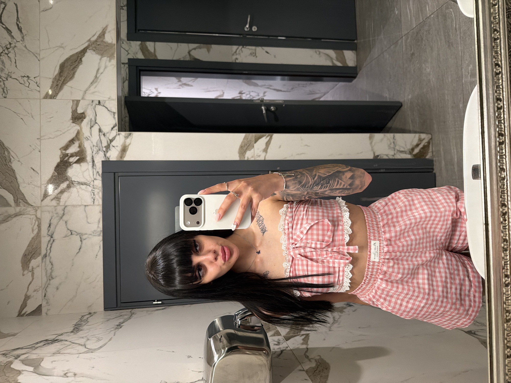 A woman with long black hair and bangs takes a mirror selfie in a public restroom. She is wearing a pink and white checkered tube top and matching shorts. She has tattoos on her arms and chest. The restroom has marble-tiled walls and dark gray doors. A hand dryer is visible to the left of the woman. The scene appears to be a modern, well-maintained public restroom, possibly in a shopping center or a public building in Plovdiv, Bulgaria.