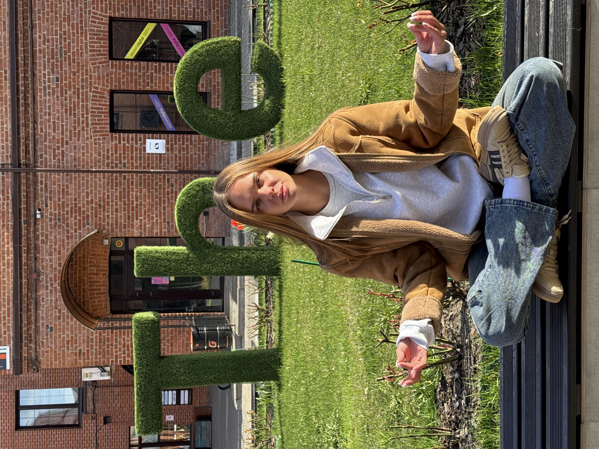 The image shows a young woman sitting cross-legged on a bench outdoors, in what appears to be a park-like setting. She is dressed in a light gray sweatshirt, a brown fleece-lined jacket, blue jeans, white socks, and beige sneakers. The woman is looking at the camera, with her arms outstretched, each hand holding a small twig.
Behind her, there is a large, topiary-like display spelling the word "GEO" in green letters, likely made of artificial turf or similar material. In the background, a brick building is visible, with various windows, including some with colored window coverings featuring "nusr/.digital" inscriptions. The location is identified through the text on a bench to be "Moscow", Russia.
The surroundings consist of green grass and some newly planted shrubs. The bench is made of dark slats. The overall lighting suggests it is daytime, with natural light illuminating the scene.