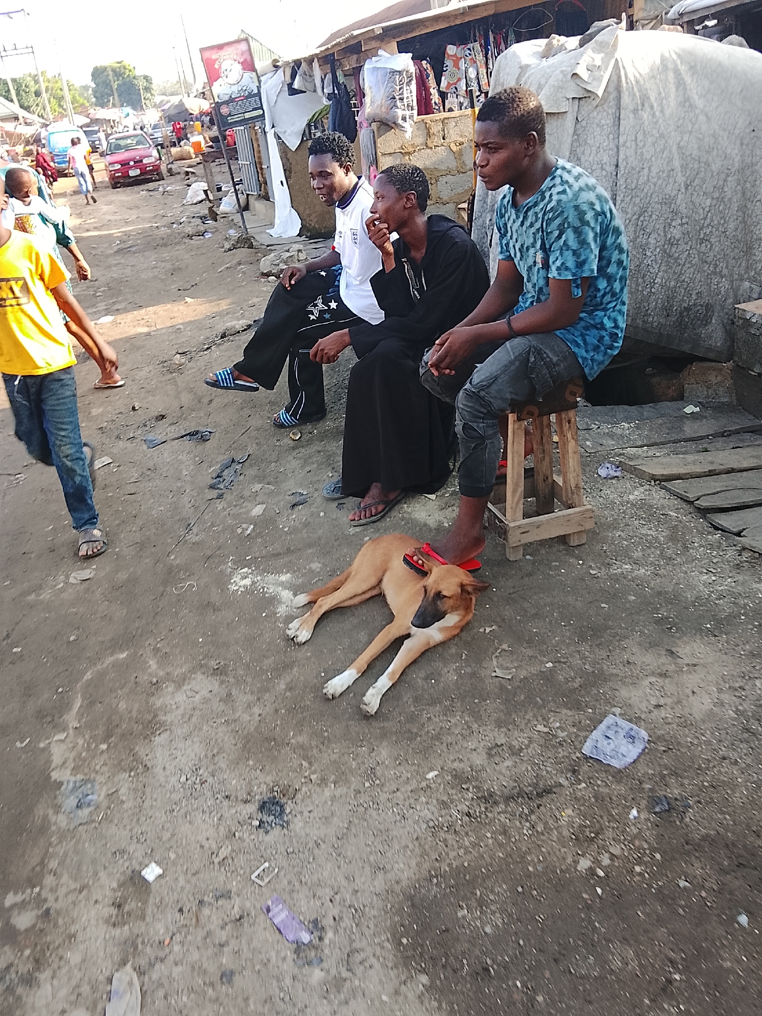 Three people are sitting along the side of a dirt road in a bustling area. One is seated on a wooden stool, wearing a blue t-shirt and jeans. The other two are sitting on what appears to be a structure behind them. One of the people is wearing a white shirt, and the other is wearing a black robe. A dog is lying on the ground near the seated people, with the person on the stool resting their foot on the dog. There are other people visible on the other side of the road as well as several vehicles. Some of the buildings on the side of the road are covered in tarps or tarpaulins. The road is dirt, and litter is scattered on the ground. Based on the environment and the clothing of the people, it appears to be located in Abuja, Nigeria.