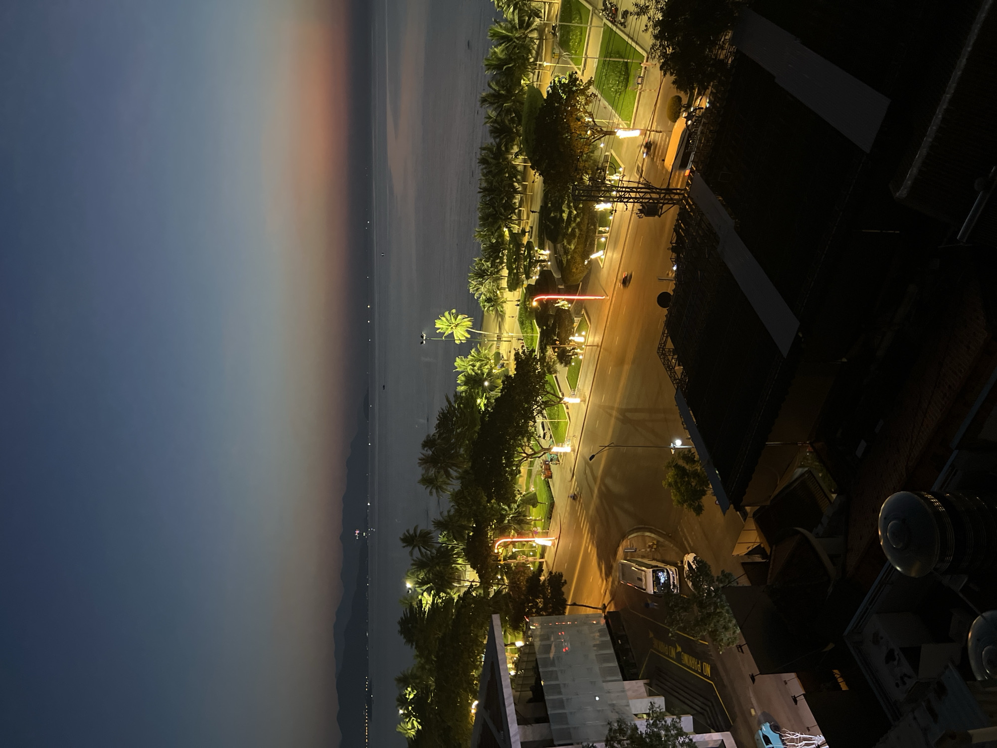The image captures a nighttime view of a coastal city, likely Nha Trang, Vietnam, characterized by a beach, a road lined with palm trees and streetlights, and distant mountains. The sky exhibits a gradient of dark blue at the top transitioning to a pale orange near the horizon, suggesting either dawn or dusk. In the foreground, a building with a modern facade and glass windows is visible, with a road in front of it. Several vehicles, including a white van and a car, are present on the road. A park area with manicured green spaces, trees, and decorative lighting is situated between the road and the beach. The beach itself is dark, with scattered lights indicating possibly distant structures or boats. The presence of palm trees, the ocean, and the general architectural style are consistent with a location like Nha Trang. There are no discernible individuals engaged in specific activities, but a few figures might be present in the park area or on the beach, though they are not clearly identifiable. The overall scene depicts a tranquil urban coastal environment at twilight.