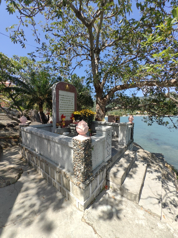 A monument stands in a seaside setting, shaded by a large tree. The structure is made of gray stone, decorated with pebble accents, lotus sculptures, and engraved figures. It sits atop a tiered platform accessed by steps. Bouquets of yellow and red flowers adorn the monument's central plaque, which is inscribed with text. The backdrop includes ocean waters, a distant shoreline, and a clear blue sky with scattered clouds. Shadows of the tree fall across the concrete foreground.
