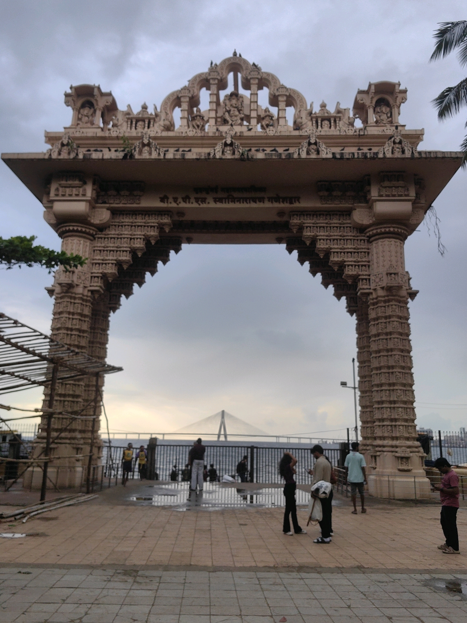 The image depicts the ornate gateway of the Shri B.R.P.S. Swami Narayan Mandir in Kalina, Mumbai. The gateway is a grand structure with intricate carvings and sculptures. Through the archway, a body of water is visible, with a bridge in the distance. Several people are present on the paved area in front of the gateway, some standing, some sitting, and some interacting with each other. The sky is overcast, suggesting a cloudy day. The overall scene is one of a religious landmark in a bustling urban environment.