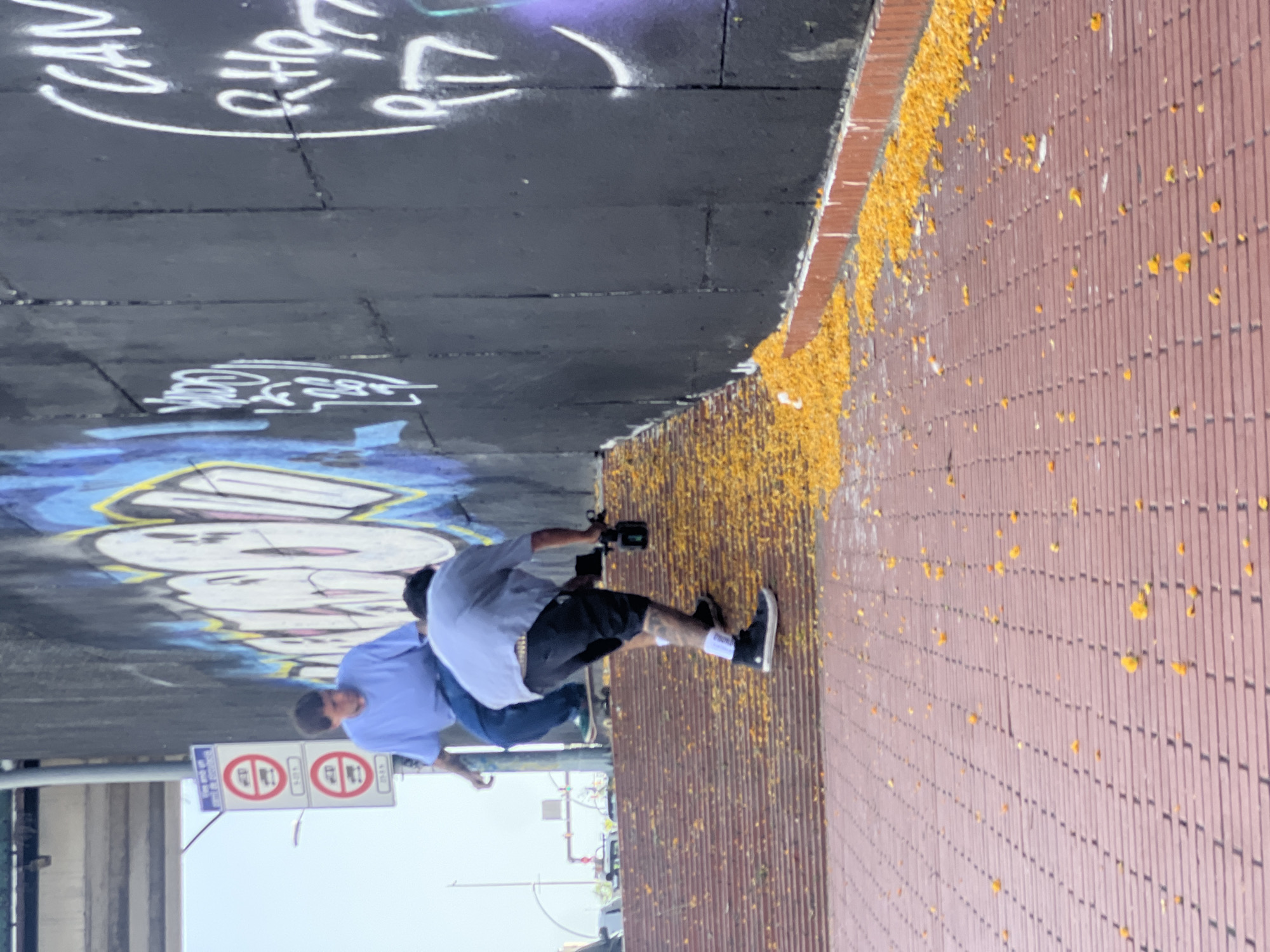 The image shows a street scene with graffiti on a wall and two people. One person is wearing a light blue shirt and jeans, and the other is wearing a grey t-shirt, black shorts, and white socks. The person in the grey t-shirt is holding a video camera and appears to be filming. The scene is outdoors, with a brick-patterned ground covered in yellow debris. Graffiti is visible on the dark wall, including stylized lettering and cartoonish drawings. Two traffic signs with red circles and diagonal lines, indicating prohibition, are also visible. The lighting suggests it might be daytime.