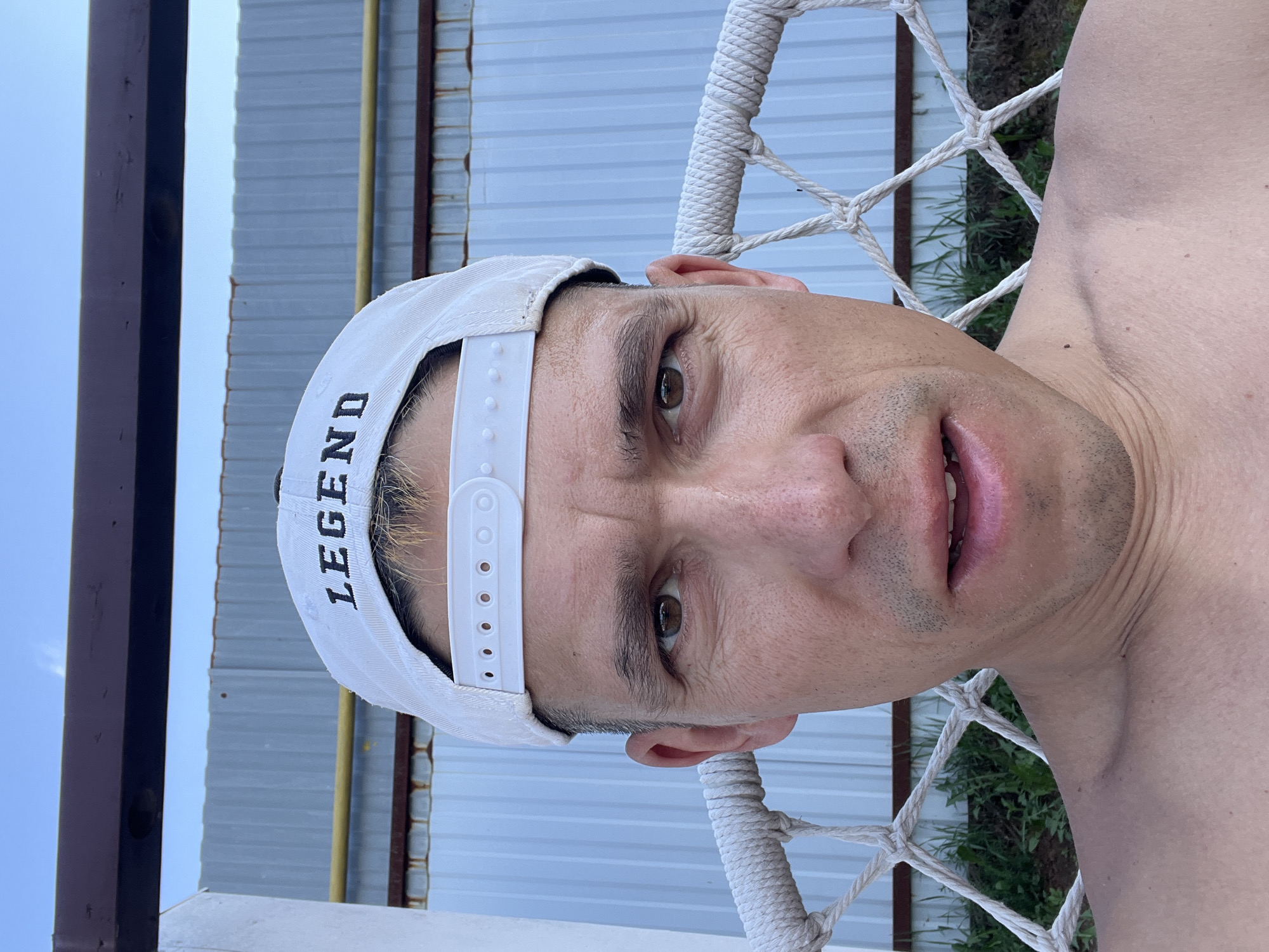 A man wearing a white "LEGEND" baseball cap is positioned in a rope swing. The setting is outdoors. He is near a metal fence. The location is Chaikovsky, Russia.