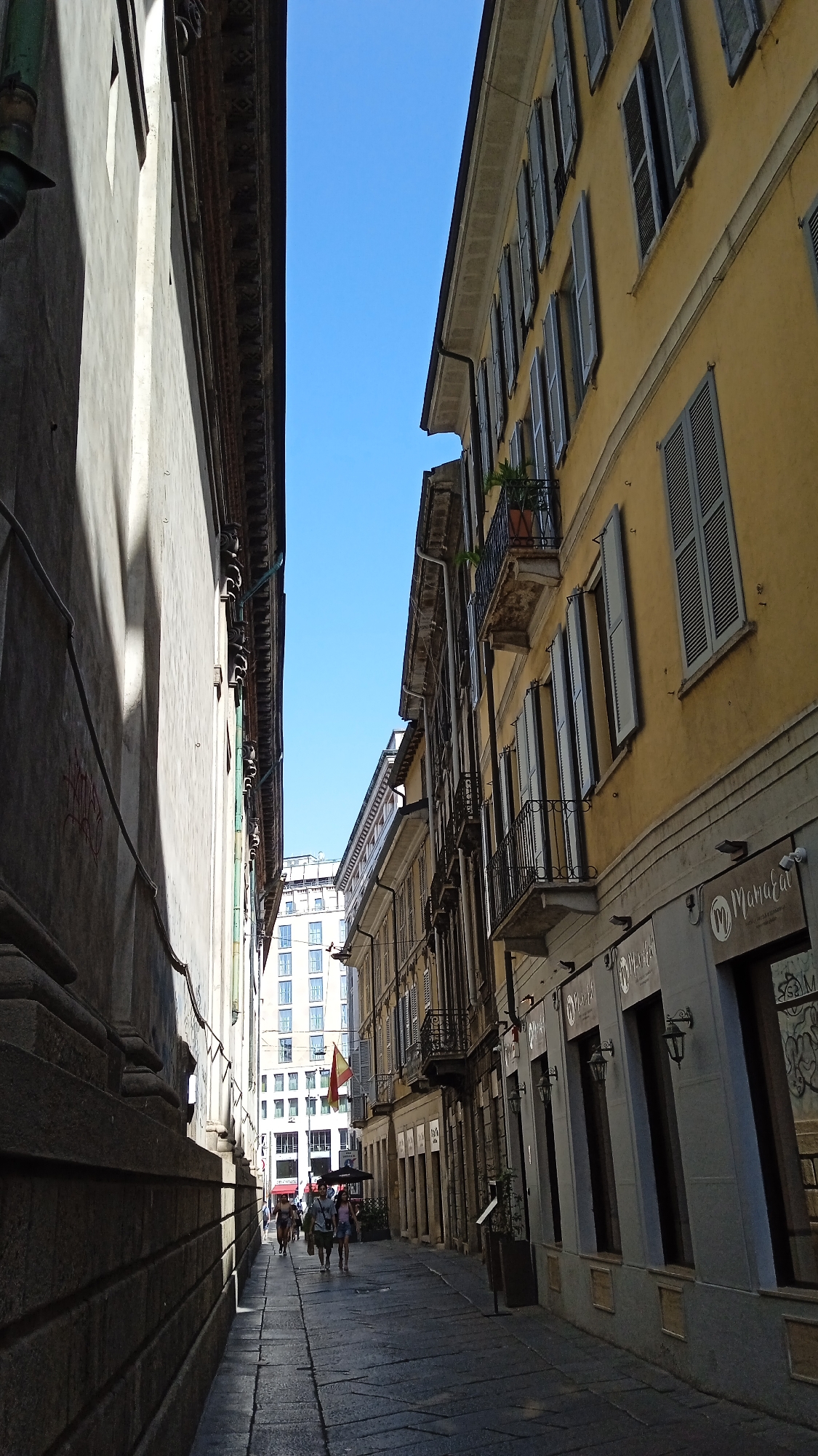 The image depicts a narrow street in Milan, Italy, flanked by tall buildings on both sides. The building on the right is painted yellow and features multiple stories with rows of open shutters. Balconies with decorative iron railings are visible. The building on the left is a muted gray color with visible stonework at its base. The street is paved with dark cobblestones. In the distance, a modern white building with many windows is visible. Several people are walking down the street, some under umbrellas, and a flag with red and yellow stripes is displayed from one of the buildings. Shops with awnings are present along the right side of the street. The sky is a clear, bright blue. The scene appears to be illuminated by strong sunlight, casting distinct shadows.
