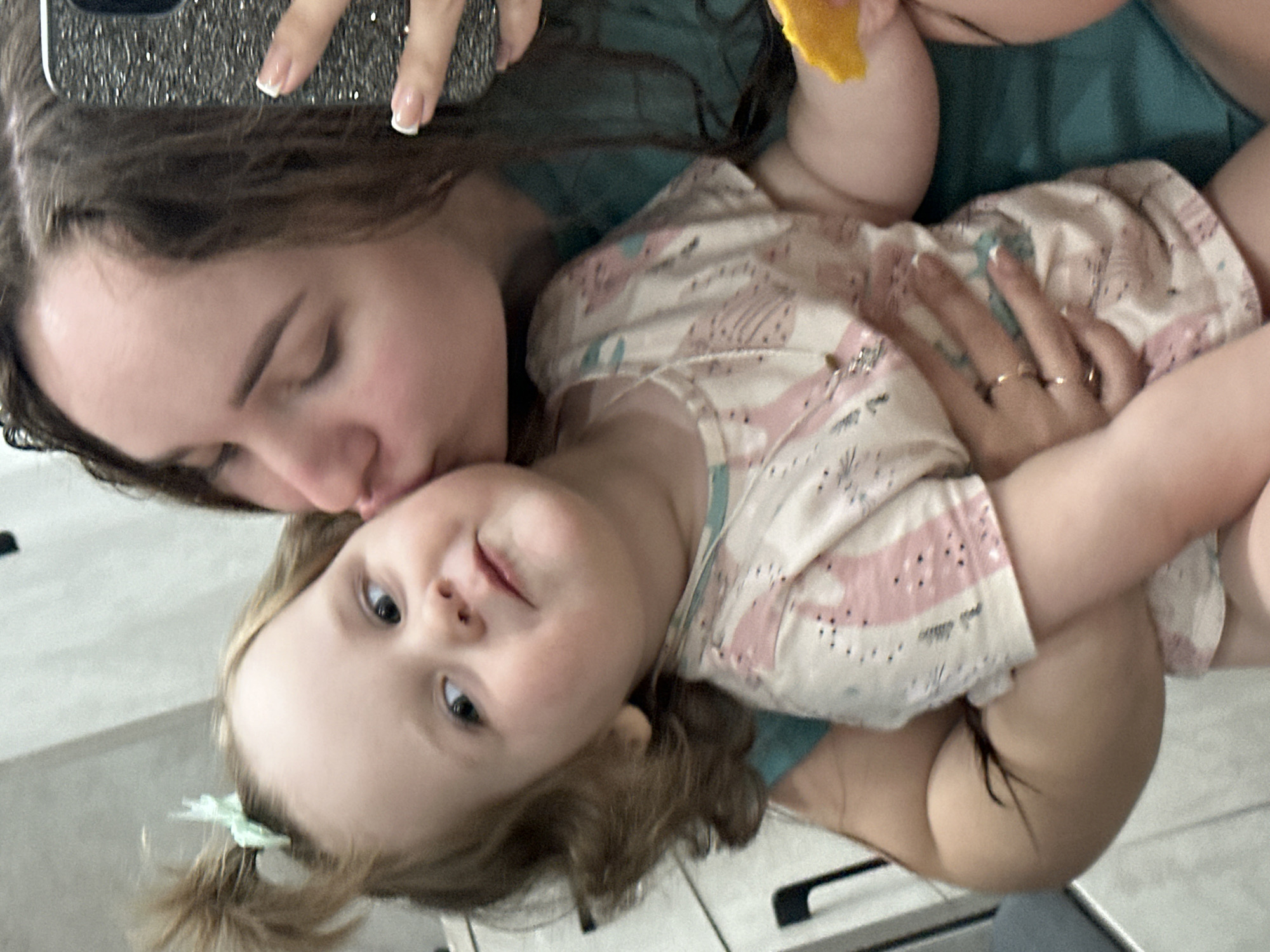 A young woman is holding a toddler. The woman has long brown hair and is kissing the toddler's head. The toddler has blonde hair styled in pigtails with a green hair accessory. The child is wearing a light-colored patterned top. The woman has a phone in her hand. The photo appears to be taken indoors with a light-colored background and possibly a counter visible. There is no confirmed evidence to determine the exact location in the city of Chaikovsky, Russia.