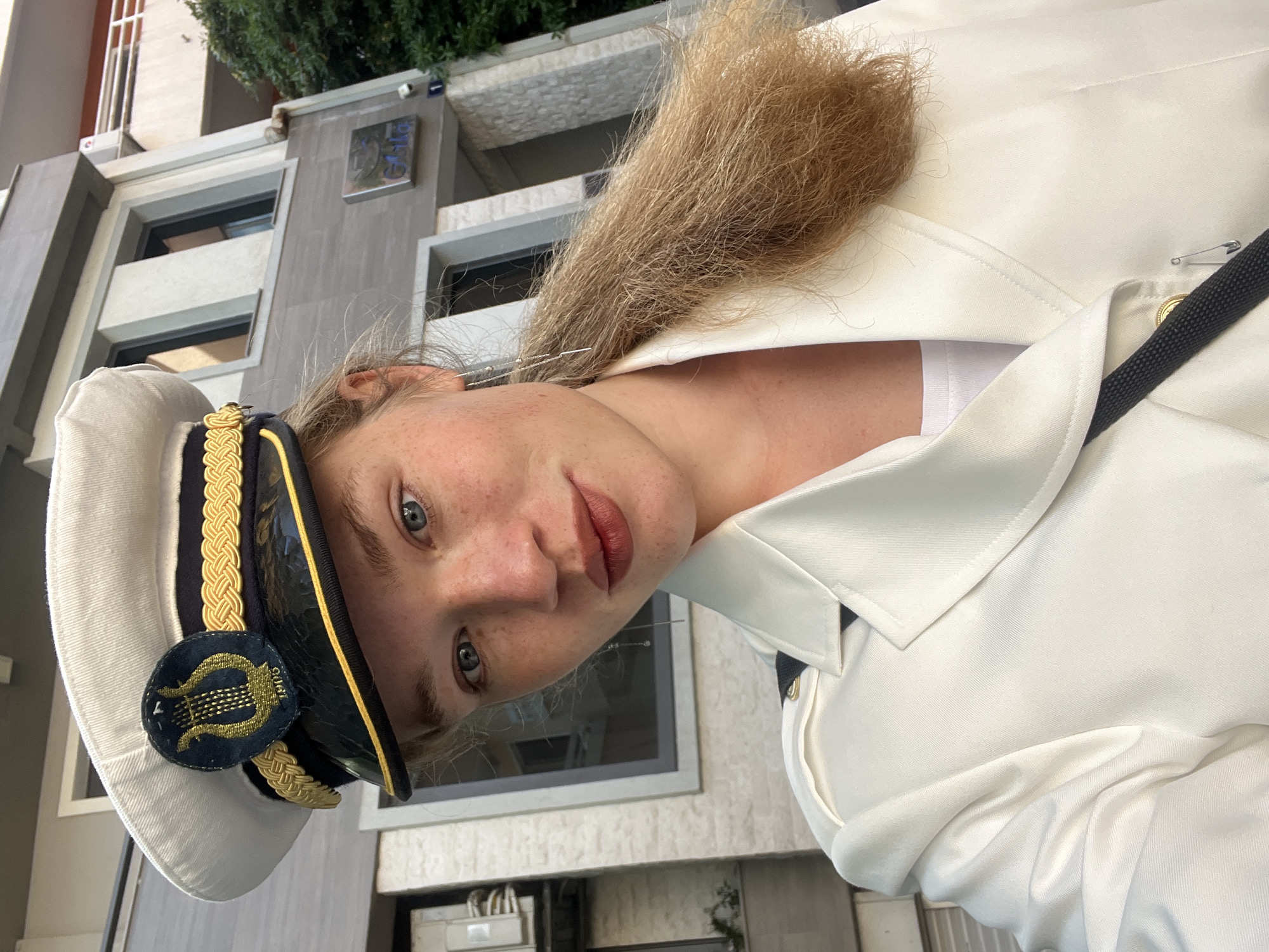 A woman with light skin and freckles is wearing a white uniform jacket and a white sailor-style hat with gold trim and a black badge featuring a harp. Her hair is blonde and appears somewhat frizzy. She is standing in front of a building with a grey facade, black-framed windows, and green foliage visible above. A plaque with a "G16" logo is visible on the building's facade. The location appears to be an urban setting, most likely Budva, Montenegro.