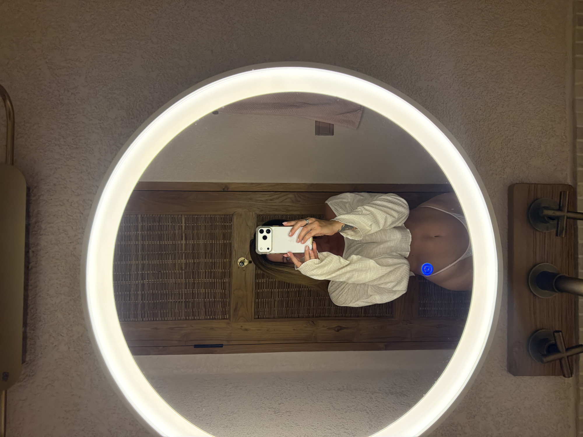A woman is taking a selfie in a bathroom mirror. The mirror is round with a built-in light. The bathroom has light-colored walls and a wooden cabinet behind the mirror. The woman is wearing a white shirt and underwear. The location is Mengwi, Indonesia.