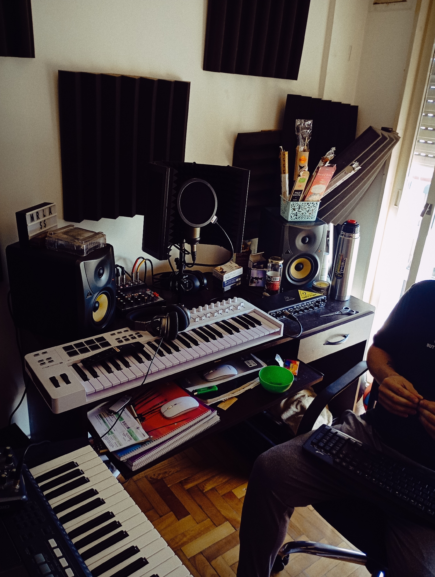 This image captures a home music studio or creative workspace during what appears to be daytime, with light entering from a window on the right. The room is equipped for audio production and recording, featuring acoustic foam panels mounted on the walls for sound treatment.
In the foreground, a person is partially visible, sitting in a black office chair. They are wearing a dark t-shirt with "BUT..." partially visible text and grey sweatpants, with a computer keyboard resting on their lap. Their hands are clasped.
The desk area is cluttered with various equipment and personal items. On the left, a studio monitor speaker with a distinct yellow cone is visible, next to a small audio mixer with numerous knobs and cables. Above the speaker, a small light box spells out "MRLA SS" and a cassette tape case rests on top. A condenser microphone with a pop filter is positioned behind the mixer, suggesting recording capabilities. Over-ear headphones are draped over a white MIDI keyboard, which occupies the central part of the desk. Below this, another larger white MIDI keyboard is also visible.
To the right of the desk setup, another studio monitor speaker matches the left one. Beside it, a silver thermos bottle and a light blue basket filled with incense sticks and packages (one clearly marked "PALO SANTO") are present. A small green bowl, a white computer mouse resting on a colorful spiral notebook, and various papers and pens are scattered on a lower shelf beneath the main desk surface. Text such as "KRK" on the speaker cone and "BEHRINGER" on an audio interface are faintly visible. The flooring is made of wooden parquet. The overall scene suggests a dedicated space for music creation or audio work.