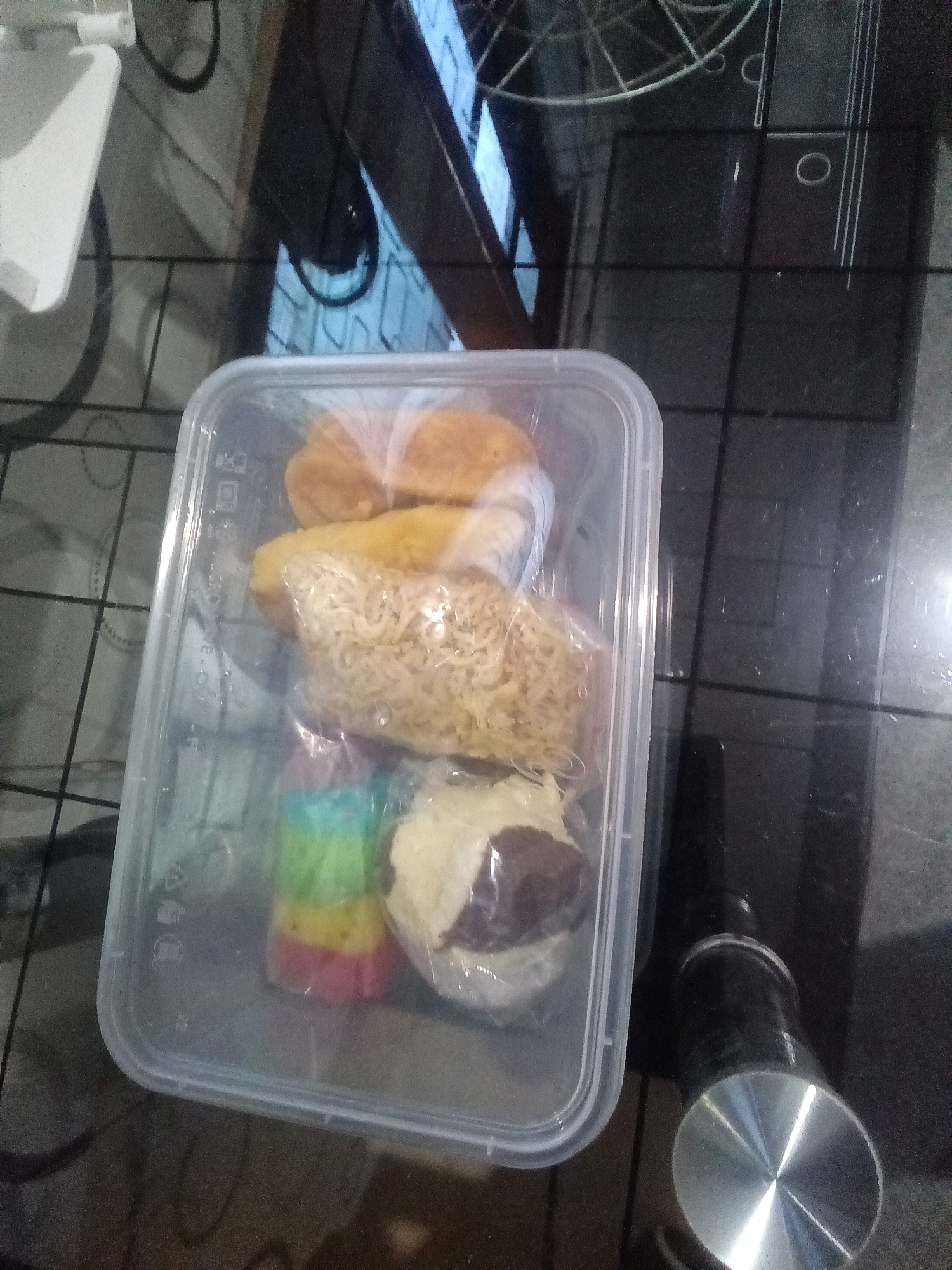 In Tegalgede, Indonesia, a clear plastic food container is shown on a dark, reflective surface, possibly a table. The container holds several food items. At the top, there appear to be fried snacks, possibly donuts or breaded items. Below them, there is a portion of dried instant noodles. Further down, a stack of colorful layers, resembling a rainbow dessert or candy, is visible. At the bottom of the container, there's a portion of what looks like a pastry or cake, possibly with chocolate filling. The overall scene suggests a takeaway meal or homemade snacks presented in a disposable container.