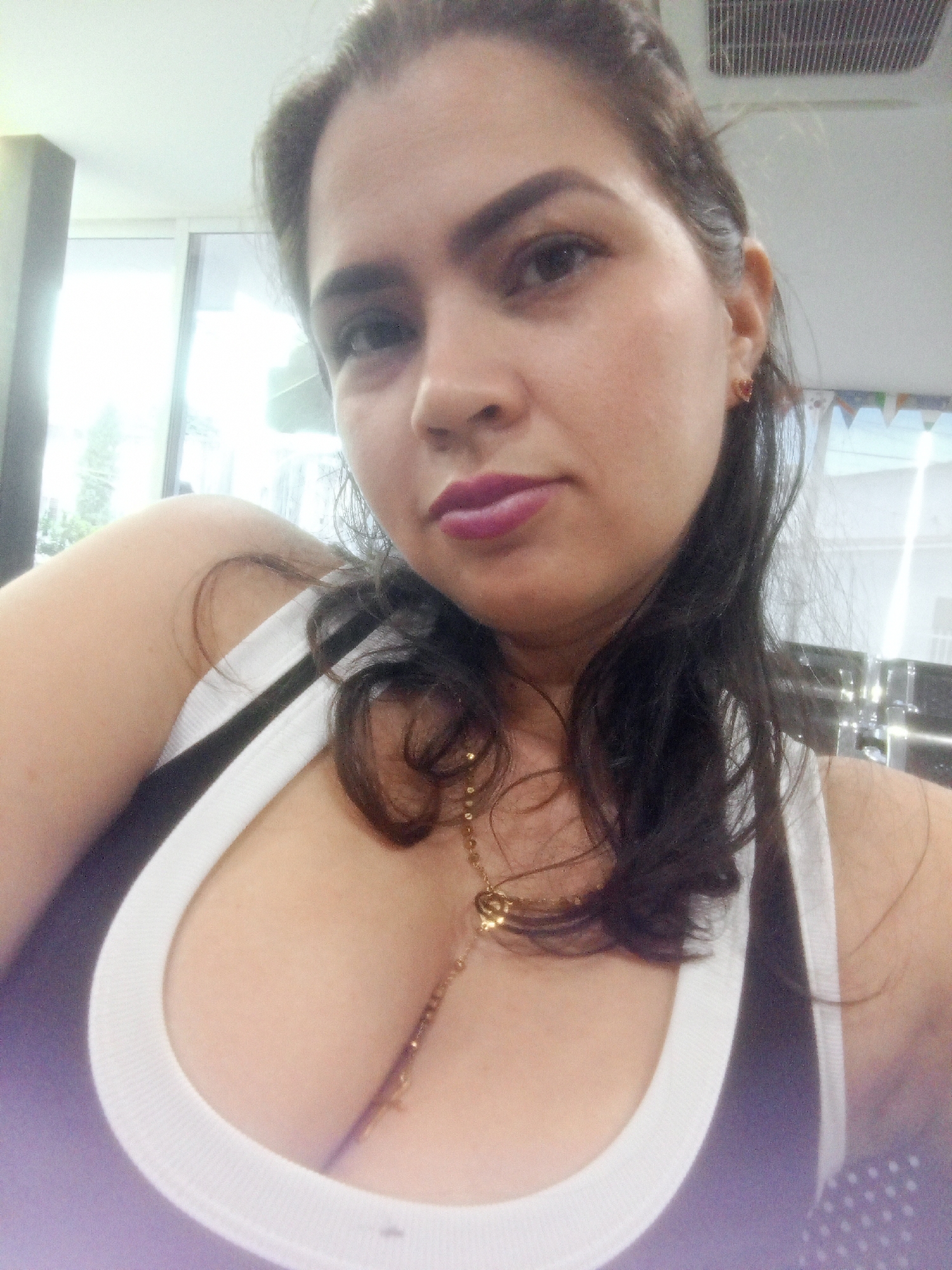 A medium shot shows a woman from the chest up, looking directly at the camera. She has dark hair, dark eyebrows, and is wearing dark lipstick. She is wearing a white tank top with a black stripe and a gold necklace. The background is out of focus and appears to be an indoor setting with fluorescent lighting. There are glimpses of windows and possibly office furniture. The location is Perímetro Urbano Cúcuta, Colombia.