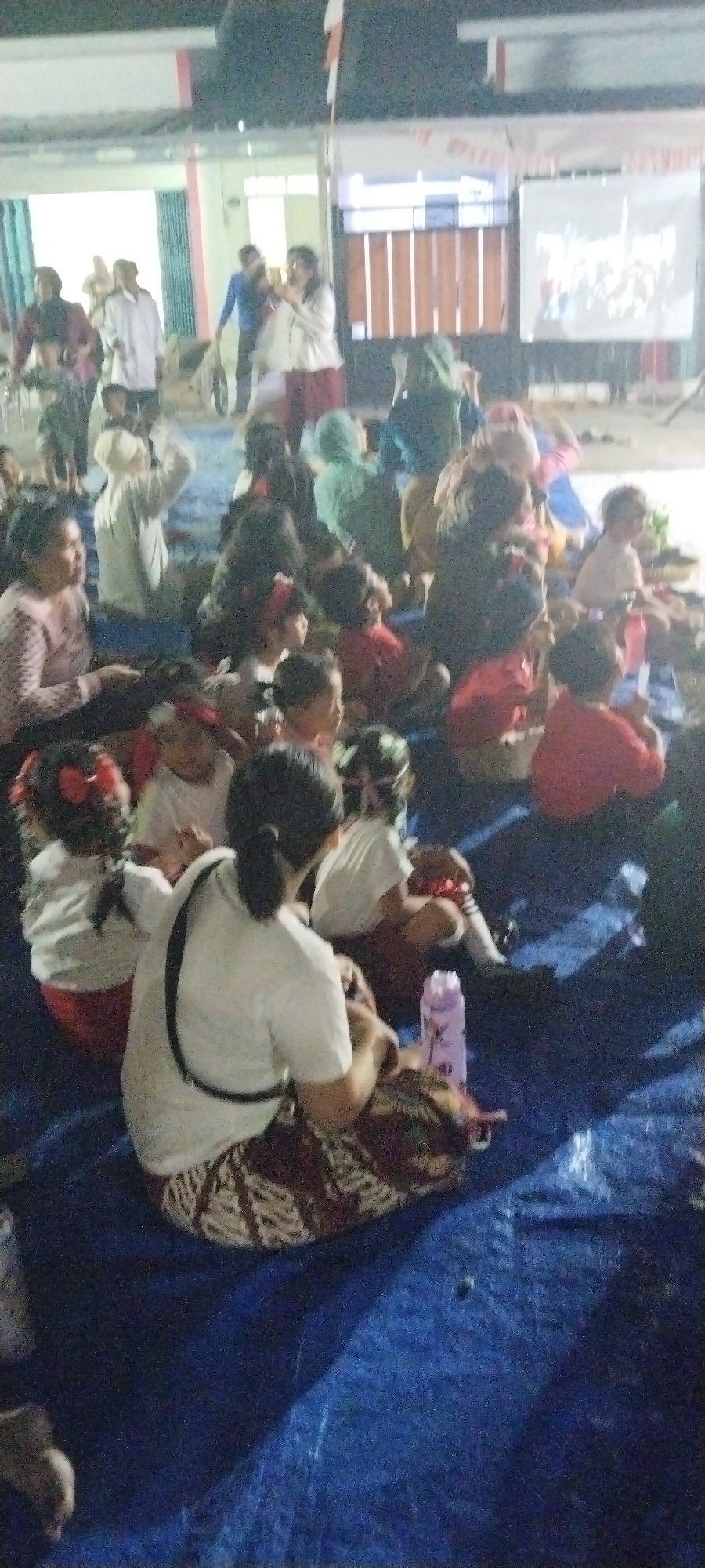 In the city of Taban, Indonesia, a group of people, predominantly children and some adults, are gathered outdoors on a large blue tarp at night. They appear to be watching a projected image on a screen in the background. The lighting suggests it's evening or nighttime. Several children are seated on the tarp, some wearing red shirts, others white. Adults are also visible, some standing and observing, while others are seated amongst the children. A woman in a white top and patterned skirt is in the foreground, facing away from the camera, with a bag strap over her shoulder. A purple water bottle is placed near her on the tarp. The environment is likely an open area, possibly a courtyard or a field, with a building structure and a fence visible behind the projected screen. The overall atmosphere suggests a communal event or gathering, possibly an outdoor movie screening or a public event.