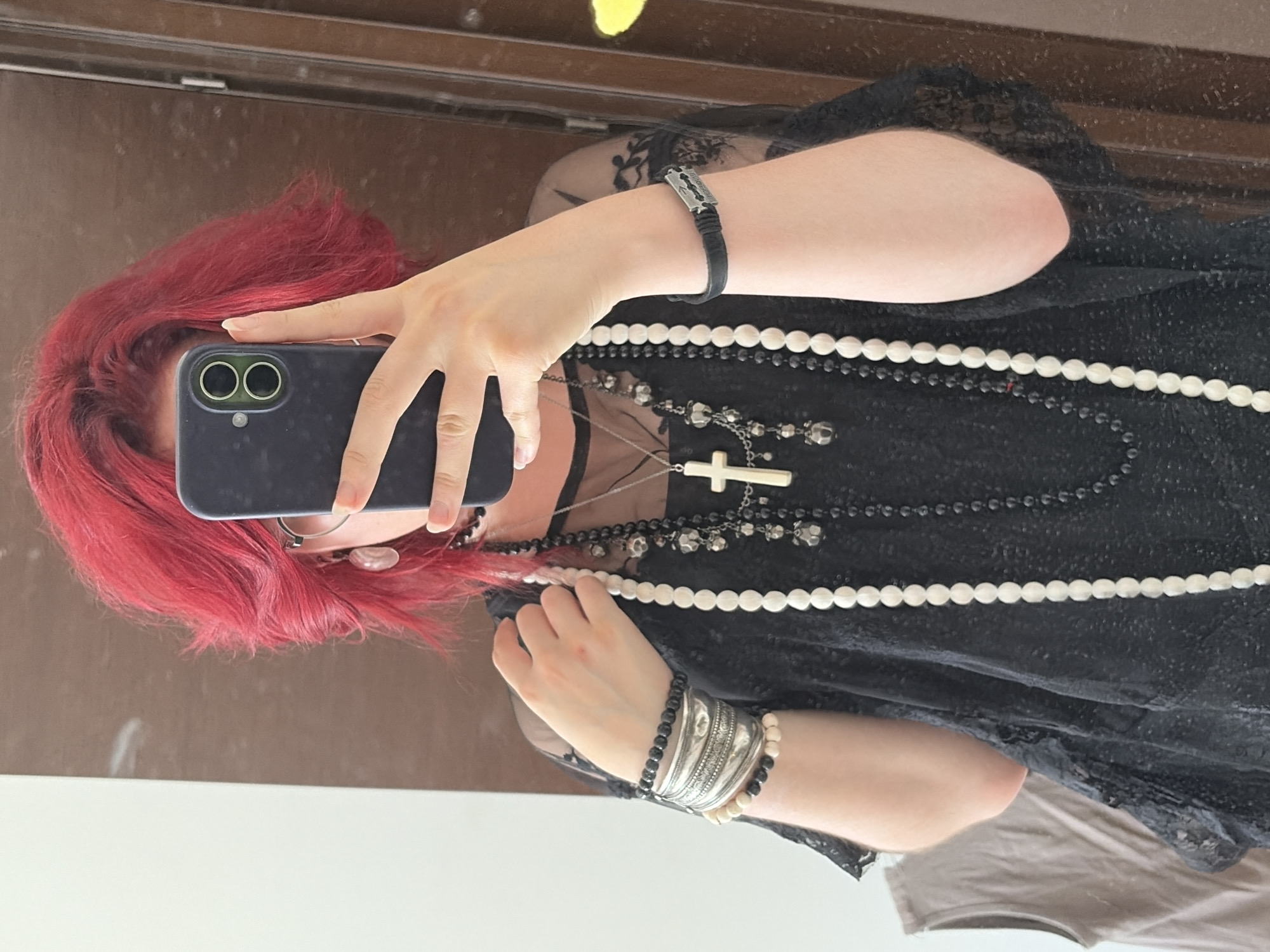 A person with bright red, choppy hair is taking a mirror selfie. They are wearing a black lace top and multiple necklaces, including one with a large white cross pendant and another with black beads and charms. They are also wearing several bracelets on their left wrist, one of which is a wide silver cuff with intricate detailing, and a black band on their right wrist with a silver accent. The person is holding a dark-colored smartphone in their right hand, partially obscuring their face. They are wearing glasses with thin, round frames. The background appears to be a room with a mirror reflecting the scene and a portion of a door frame. There is also a hint of a gray garment hanging on the left side of the frame. The image does not contain any recognizable landmarks or indicators of its specific location in Chelyabinsk, Russia.