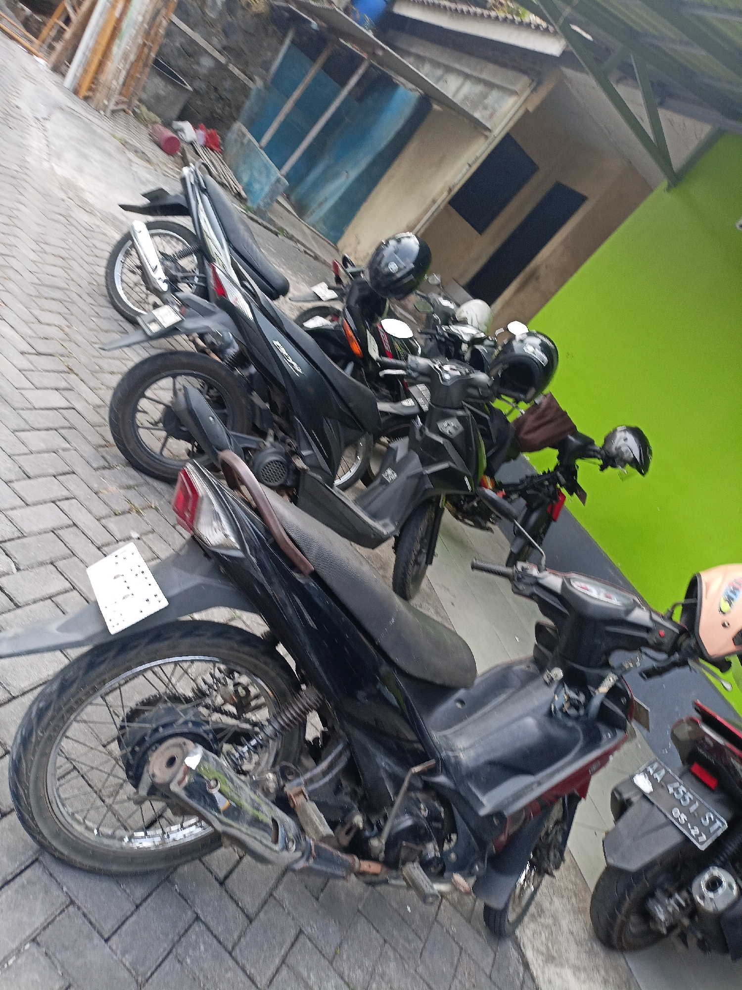 The image depicts a row of parked motorcycles in what appears to be an outdoor area in Wancen, Indonesia. The motorcycles are primarily black and are parked on a brick-patterned pavement. Several helmets are visible, some placed on the seats of the motorcycles and others on the handlebars. In the background, there is a building with a lime green wall and a section of a weathered structure with wooden elements and what looks like a blue tarp or sheeting. The scene suggests a typical streetscape where motorcycles are commonly used for transportation.