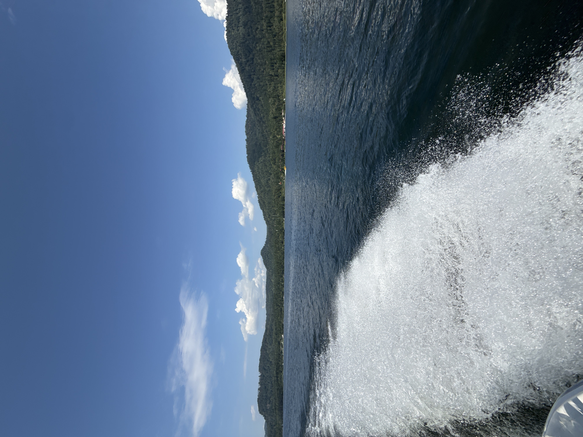 This image depicts a scene on Lake Teletskoye near Artybash, Russia. The perspective is from a moving boat, indicated by the large white wake generated on the left side of the frame. The water is a deep blue, with subtle ripples. In the distance, a densely forested mountain range forms the horizon. The sky is a vibrant blue, dotted with scattered white cumulus clouds. A small yellow boat is visible in the water near the distant shore, and buildings and structures can be seen nestled amongst the trees on the mountainside. The sun appears to be shining brightly, casting a clear light on the landscape.