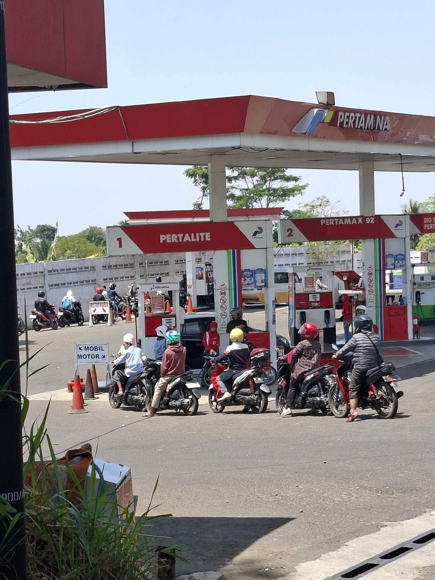 The image depicts a Pertamina gas station in Cikahuripan, Indonesia, on a sunny day. Several motorcycles and a car are present, with people refueling their vehicles. A sign indicates "PERTALITE" and "PERTAMAX 92". The gas station has a red and white color scheme, typical of Pertamina. The environment appears to be an open area with trees and a concrete wall in the background.
