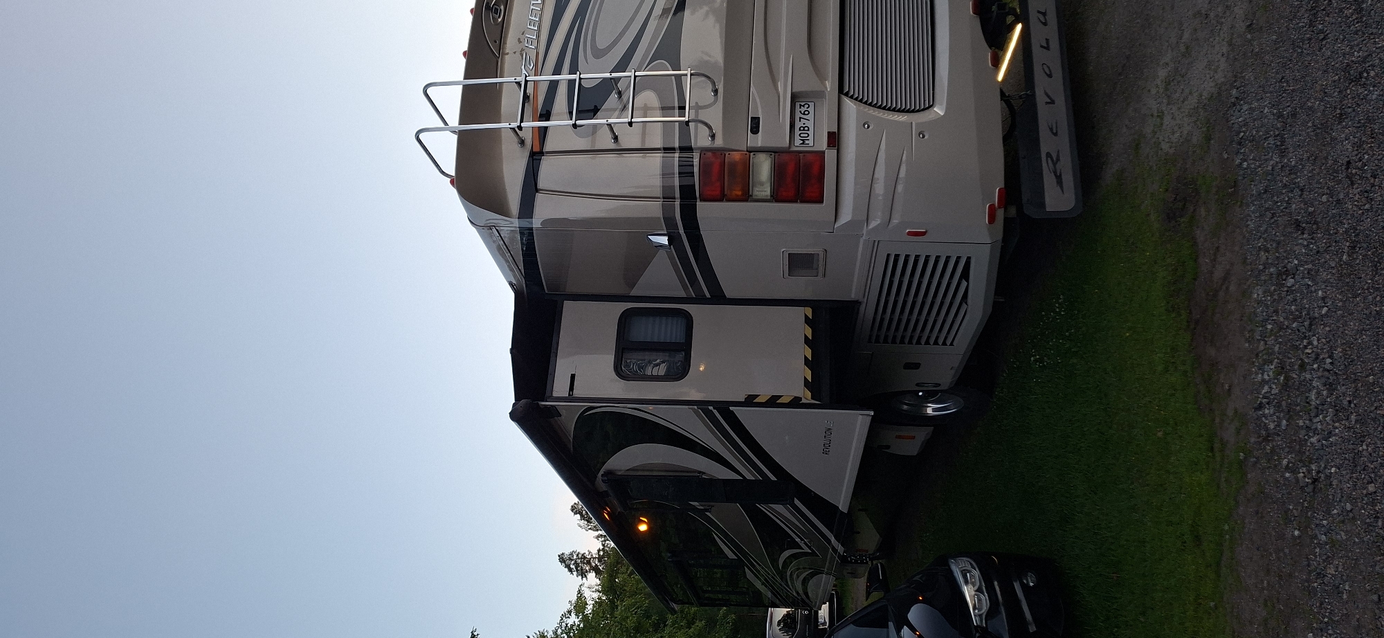 A fleetwood Revolution RV is parked on a grassy area next to a gravel path. The RV is light beige with a dark gray and black graphic design. The driver's side of the RV is visible, with the awning extended. A silver ladder is attached to the rear of the RV. A black car is parked to the left of the RV. Trees are visible in the background. The sky is clear and pale blue. The license plate on the RV reads MOB-763. The RV has a "REVOLUTION" logo on the rear. The image is taken from a low angle.