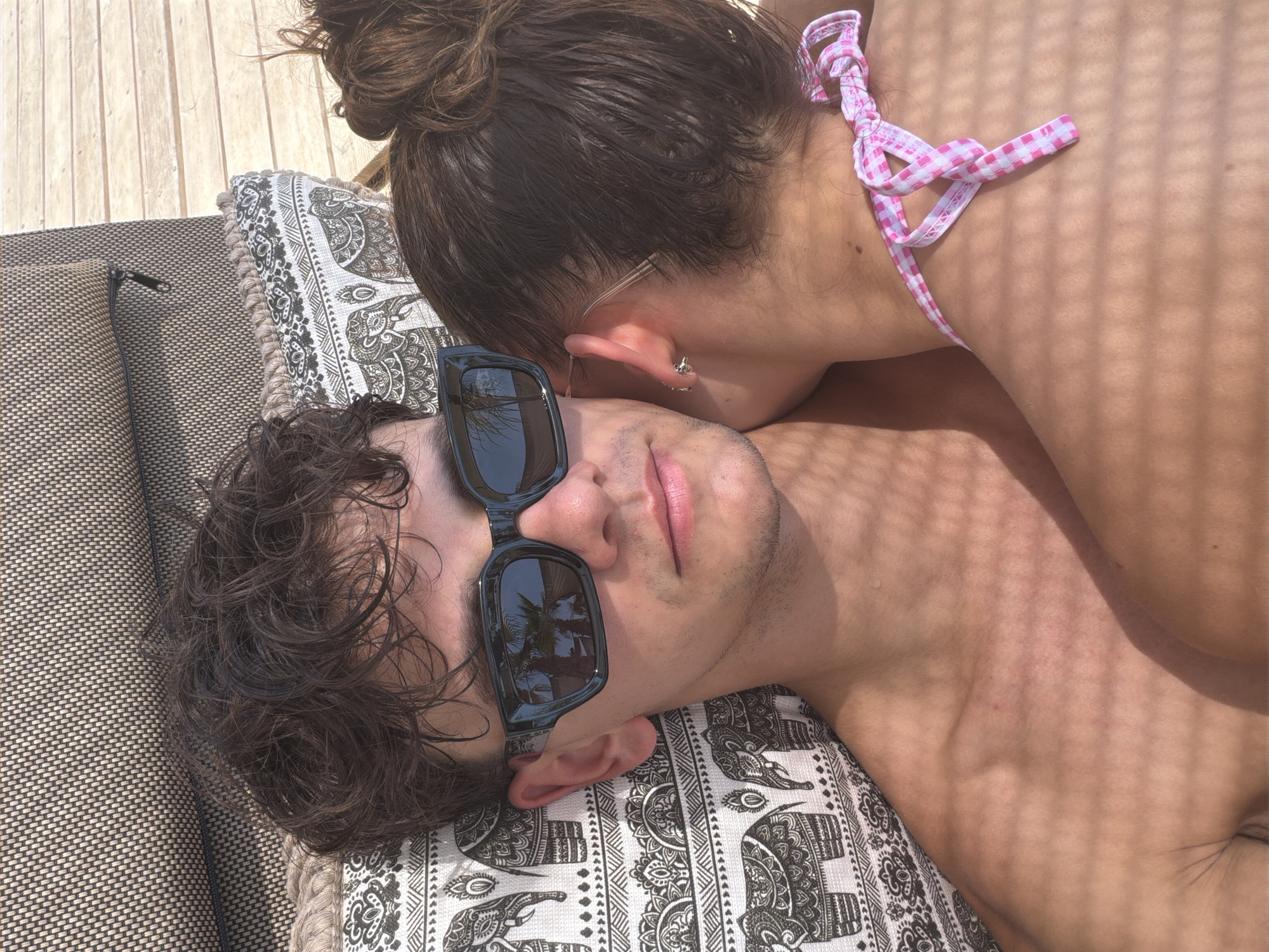 The image captures a close-up view of a young couple resting on a lounge chair at Ofrynio Beach, Greece. The man, positioned in the foreground, wears black sunglasses that reflect palm trees and the sky, suggesting a sunny, tropical environment. He has dark, tousled hair and appears relaxed. Beside him, a woman leans in, her head resting on his shoulder. Her hair is pulled back in a bun, and she wears a pink and white checkered bikini top. The background shows the patterned fabric of the lounge chair and a hint of wooden decking, indicative of an outdoor seating area common at beach resorts. The lighting suggests it is daytime, with dappled shadows falling across the subjects, likely from overhead foliage or an awning.