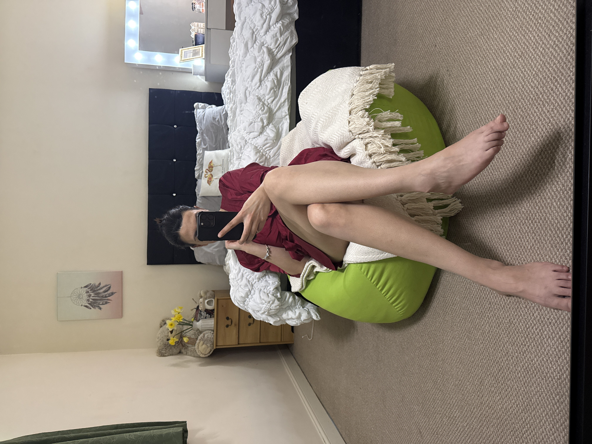 An adult individual is depicted sitting on a lime green beanbag chair, holding a black smartphone (identified as an iPhone by its camera array) towards themselves, indicating a mirror selfie. The individual's face is obscured by the phone. They are wearing a dark red/maroon robe or dress, and a silver-colored bracelet is visible on the left wrist. Their bare legs are crossed at the ankles and extended forward, with bare feet visible on the carpet. A cream-colored throw blanket with tassels covers part of the beanbag chair.
The scene is an indoor bedroom. In the background, a bed with a dark, upholstered headboard featuring four diamond-shaped embellishments is visible. The bed has a white, textured duvet and grey pillows, including one decorative pillow with a flamingo design. To the left of the bed is a light brown wooden chest of three drawers, on which a light brown teddy bear, a white vase containing yellow daffodils, and a small animal figurine are placed. A rectangular canvas print featuring a dreamcatcher design hangs on the wall above this bedside table. Dark green patterned curtains are visible on the far left. To the right of the bed, a white/light grey vanity table with a rectangular mirror bordered by exposed light bulbs is present. On the vanity, a framed picture, a white object (possibly a candle or small vase), and multiple small bottles (likely perfume) are arranged. The room features light-colored walls and a light brown/beige carpet with a textured pattern.
The image does not contain any visual information to specify a location within Great Yarmouth, United Kingdom, beyond being an indoor bedroom.