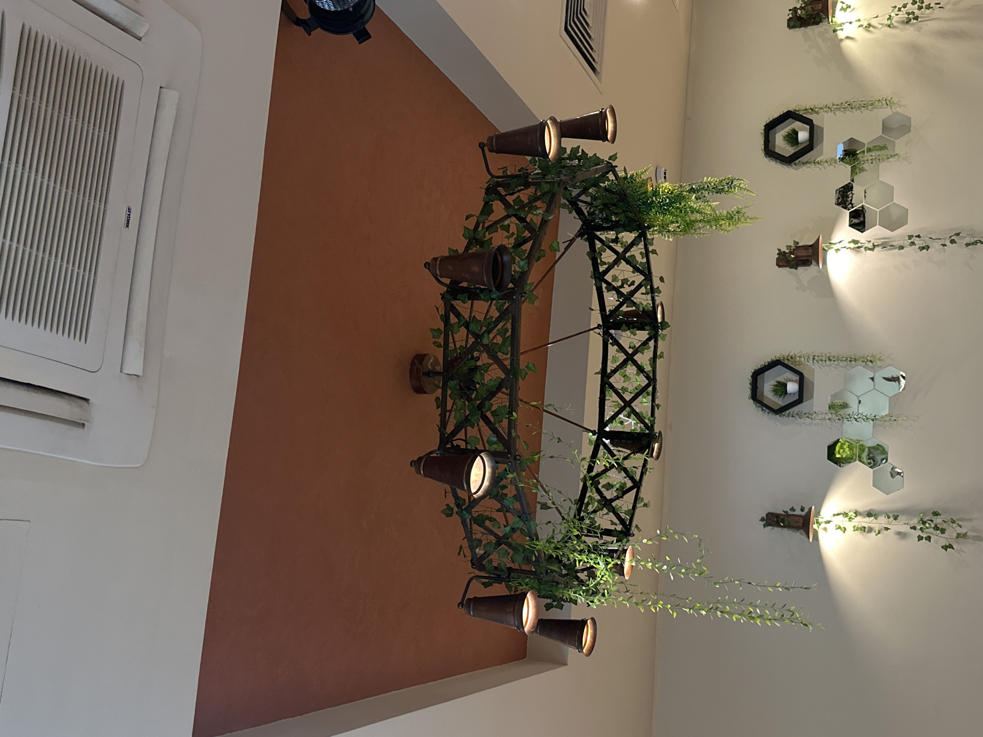 An indoor scene shows a ceiling with a decorative chandelier and an air conditioning unit. The chandelier is circular with a dark metal frame interwoven with artificial green vines. It has several conical, dark-colored light fixtures hanging from it, each emitting a warm light. Hanging artificial vines also drape from the chandelier. Below the chandelier, on the wall, are several decorative elements. These include wall sconces with conical light fixtures and artificial vines, as well as hexagonal mirrors arranged in a cluster. Some of the mirrors reflect the artificial plants. A hexagonal shelf with a small potted plant is also visible. The walls are painted a light color, and the ceiling is a mix of white and a warm, reddish-brown hue. The air conditioning unit on the ceiling is white and has visible vents. This image was taken in Sochi, Russia, at a restaurant called "Chekhov's Garden."