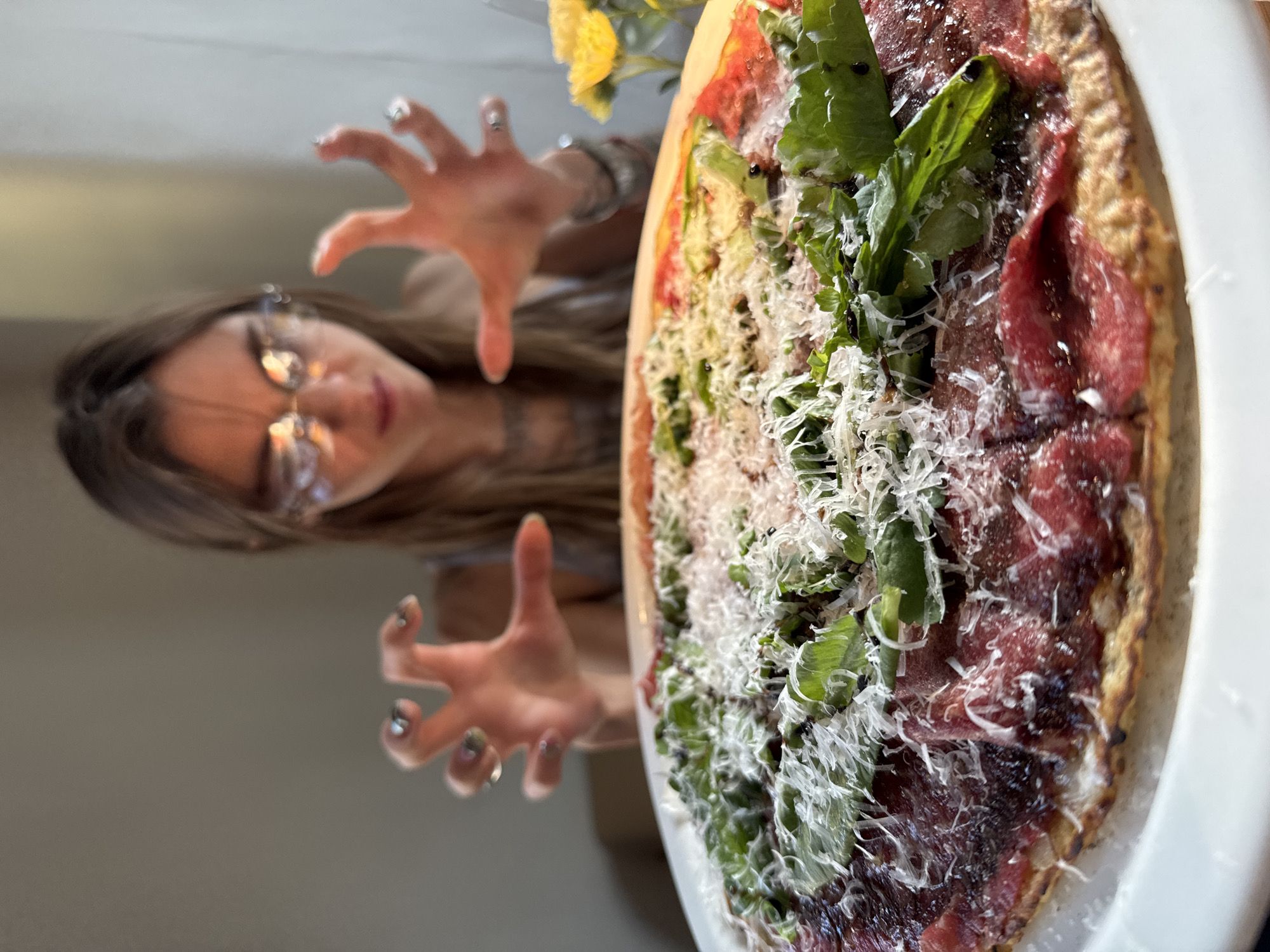 An intimate, low-angle photograph captures a gourmet pizza in the foreground, in sharp focus, with a woman partially visible in the blurred background. The pizza, served on a white plate, is generously topped with thin slices of red meat (possibly bresaola or pastrami), fresh green arugula leaves, and a substantial amount of grated white cheese, all resting on a crisp, golden-brown crust.
In the background, a woman with long brown hair and glasses is positioned to the left. She extends both hands towards the pizza, fingers splayed in a playful, almost claw-like gesture, indicating eagerness or anticipation. Her fingernails have a distinctive shiny, metallic polish. Her face is slightly out of focus, but her gaze appears directed at the food. The overall lighting is bright and natural, suggesting a daytime indoor setting. Faint yellow flowers are subtly visible in the upper background. The photo creates a dynamic perspective, emphasizing the deliciousness of the food and the playful interaction with it. The location context for this image is Moscow, Russia.