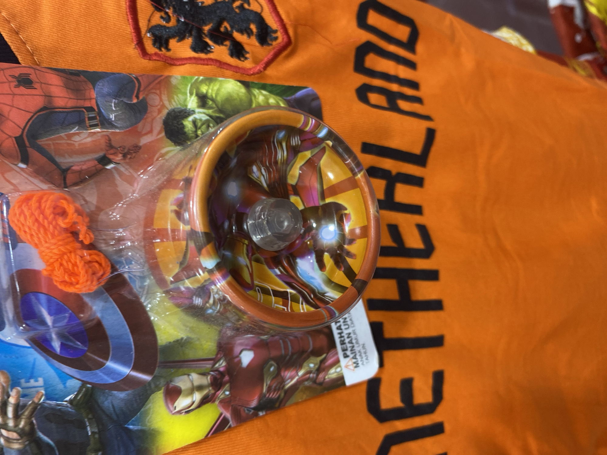 This is a close-up, eye-level, indoor shot of a t-shirt with the word "NETHERLAND" printed in large black capital letters. The t-shirt is orange and has a patch on the right side depicting a black lion rampant on a red background, which is the coat of arms of the Netherlands. In the background, to the left, is a retail display of Marvel superhero merchandise. Specifically visible are Captain America's shield, Iron Man, Spider-Man, and the Hulk. A transparent plastic package contains two orange yo-yos. The merchandise appears to be for sale. The scene is likely in a shop or market in Banda Aceh, Indonesia, given the multilingual text on a warning label attached to the yo-yo packaging which includes "PERHATIAN MAINAN UNTUK ANAK UMUR DI ATAS TAHUN" (Warning Toy for children over years old), indicating it is likely a local product or import for the Indonesian market. The lighting is neutral and even.