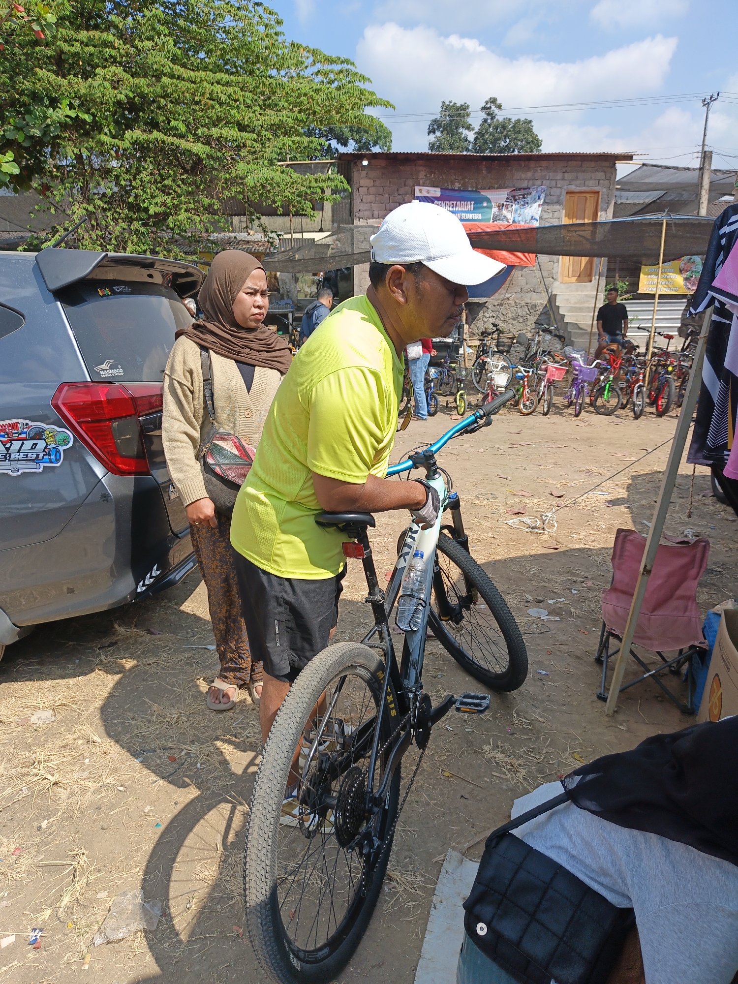 In Jatinom, Indonesia, a man wearing a bright yellow t-shirt and a white cap is standing next to a bicycle. He has his hand on the handlebars. Beside him, a woman wearing a brown hijab and a light brown cardigan stands looking away from the camera. She is wearing patterned pants and sandals. A gray car is parked to their left. In the background, a variety of bicycles are displayed under a canopy, and there are some buildings with corrugated metal roofs. The ground is dirt and covered with dry grass. The sky is blue with scattered clouds.