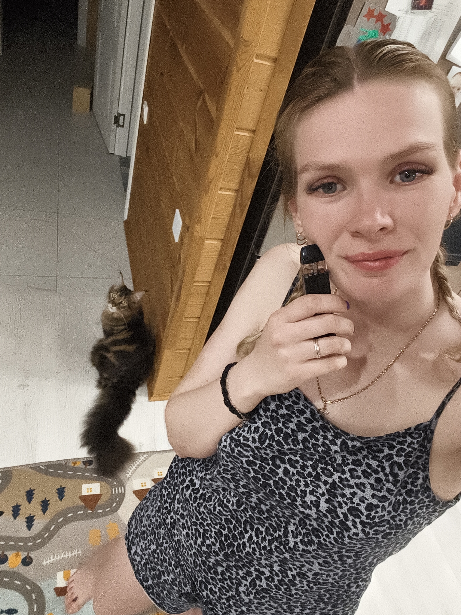 A person is holding an electronic cigarette, positioned at an angle, in an indoor setting. The person is fair-skinned with blonde hair in braids, wearing a black and grey animal print top with thin straps, and a gold necklace. On the floor is a cat sitting near a patterned rug depicting roads, vehicles, and buildings. The background features a partially visible door and a light-colored tiled floor. The location is potentially in Русский Вожой, Russia.