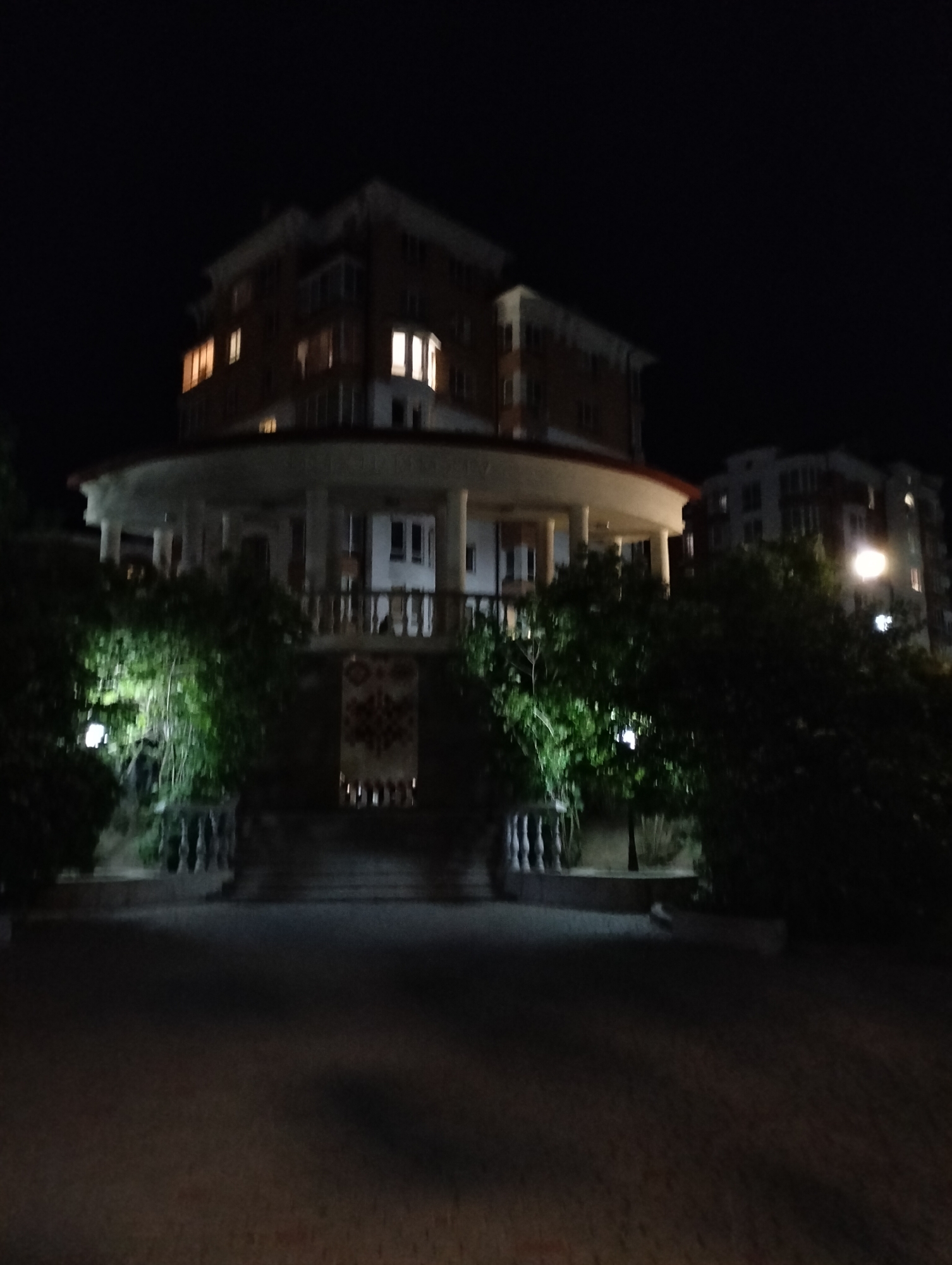 At night, the scene captures a round, gazebo-like structure with white pillars and a red roof, standing in front of a taller building with illuminated windows. The area is surrounded by dark greenery, and the ground appears to be a paved surface. The location is Krykhivtsi, Ukraine.