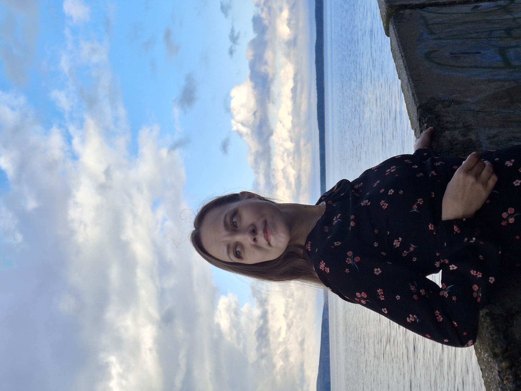 A woman is sitting on a concrete structure overlooking a body of water. She is wearing a dark dress with small red floral patterns. To her left, the sky is visible, filled with white clouds against a blue background. The body of water is a light blue-gray color. Graffiti is visible on the concrete structure on which she is sitting. The image was taken in the city of Chaikovsky, Russia.