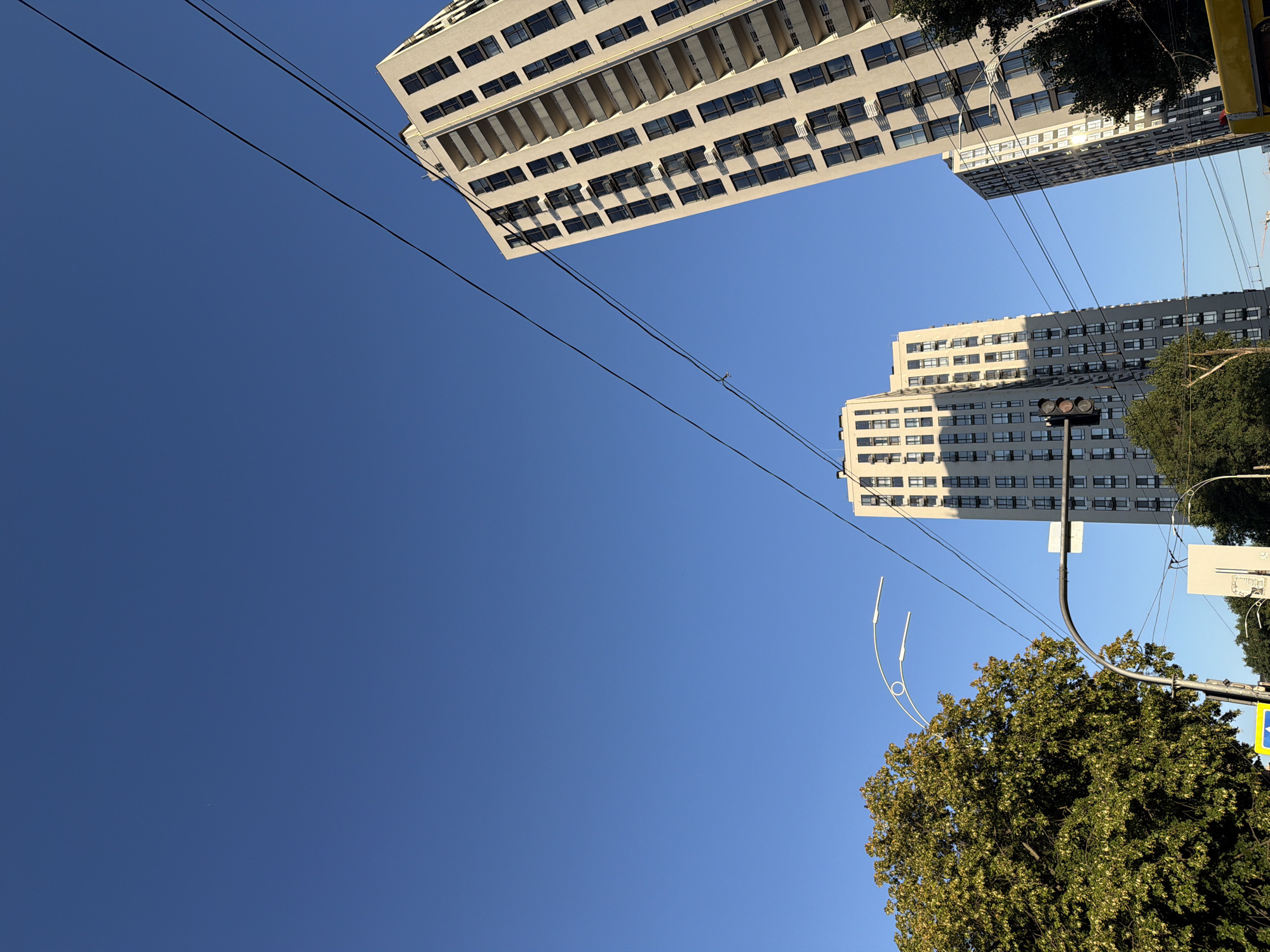 The image shows a street scene in Kyiv, Ukraine, with several modern apartment buildings under a clear blue sky. In the foreground, a large tree with green and yellow leaves is visible on the left. A traffic light is positioned to the right of the tree. In the middle ground, two tall apartment buildings stand side by side, with one being darker in color than the other. A third, taller apartment building is located to the right of these, angled towards the sky. Overhead, power lines crisscross the frame. The buildings are characterized by their uniform windows and balconies. The overall impression is of a contemporary urban environment bathed in sunlight.