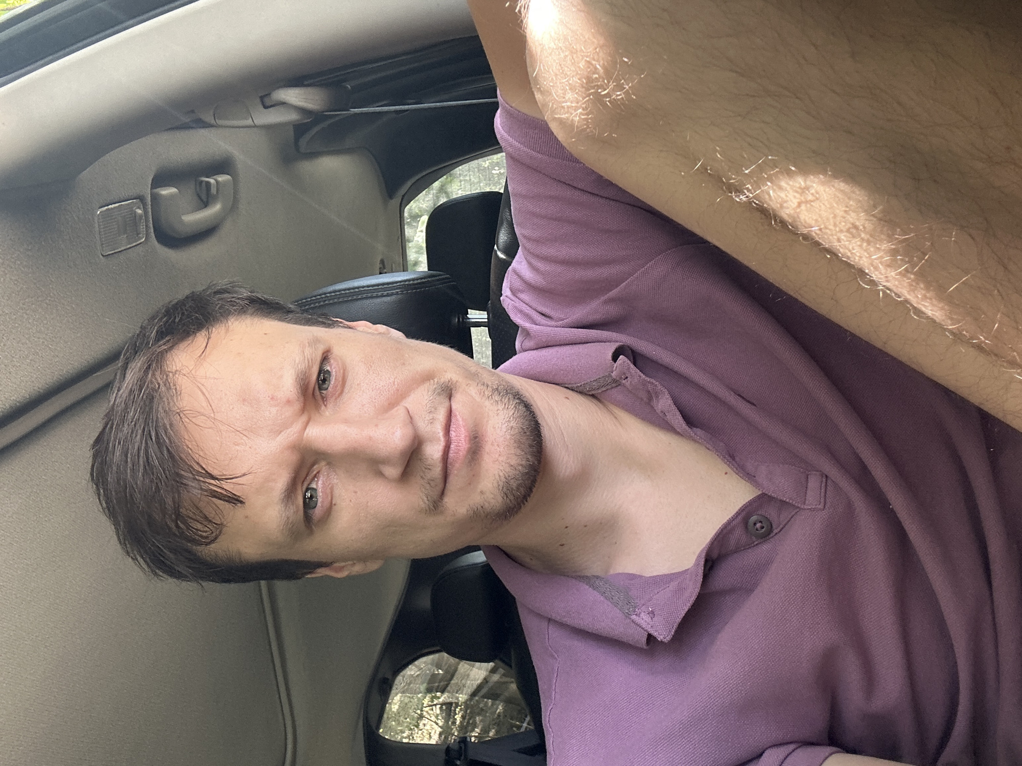 A young man is pictured from the chest up, seated in the driver's seat of a vehicle. He is wearing a short-sleeved purple polo shirt. His head is turned slightly to his left, and he is looking directly at the camera with a serious expression. Sunlight is visible on his right arm and shoulder, casting a bright highlight. The interior of the car is visible behind him, including the headrest of the passenger seat and a portion of the car's ceiling. The background outside the car window shows foliage and trees, suggesting a setting with natural surroundings, possibly on the outskirts of Makiv, Ukraine, given the context of the request.