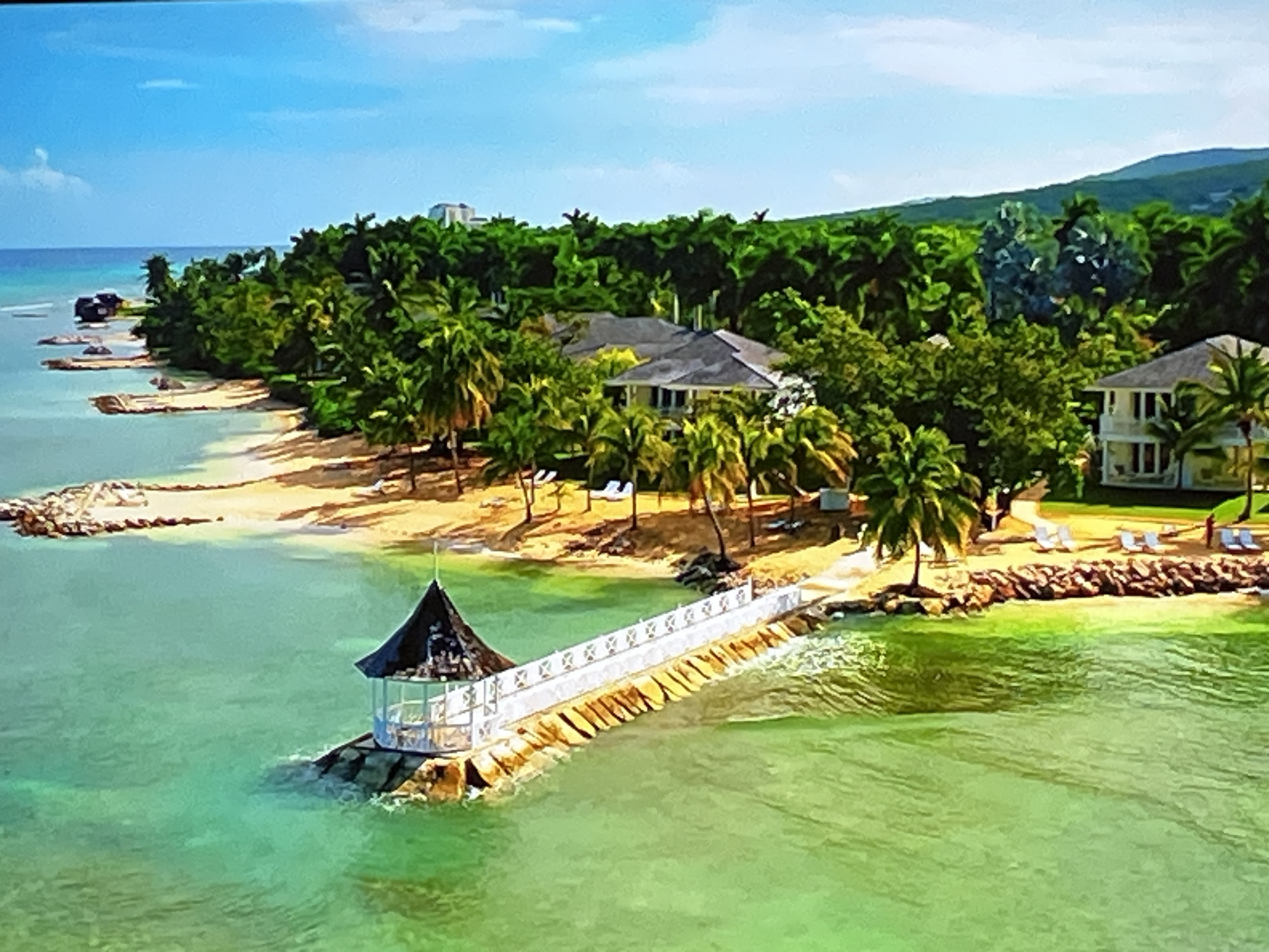 The image shows a tropical resort with a white pier extending into turquoise water. A gazebo with a dark conical roof sits at the end of the pier on a rocky outcrop. The shore is lined with palm trees and sandy beaches, with several white lounge chairs visible. In the background, there are buildings with white facades and grey roofs, nestled amongst lush green foliage. A mountainous landscape forms the backdrop, under a clear blue sky with a few wispy clouds. The scene is idyllic and suggests a vacation destination. There is no visible indication of the city of Kharkiv, Ukraine.