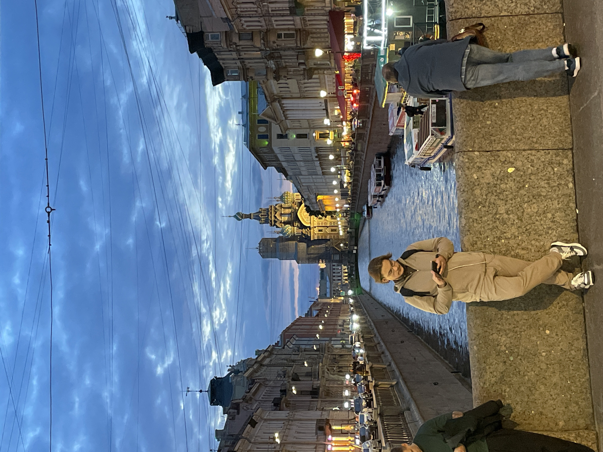 The image captures a view of the Griboyedov Canal in Saint Petersburg, Russia, at dusk. In the foreground, a young man with glasses and medium-length brown hair is leaning against a stone railing, looking at his phone. He is wearing a beige tracksuit and sneakers. To his left, another person, partially visible, is wearing a dark green shirt and holding a dark jacket. To the right of the young man, a man in a blue hoodie and jeans is leaning over the railing, facing away from the camera. He has a brown bag slung over his shoulder.
The canal is lined with buildings on both sides, characteristic of Saint Petersburg's architecture. On the left, buildings with illuminated windows and lampposts line the embankment. On the right, similar buildings are visible, with red awnings over some ground-floor establishments. Several boats are docked along the canal's edge, and a few are in motion.
In the distance, the iconic Church of the Savior on Spilled Blood stands prominently, its colorful domes illuminated against the twilight sky. The sky itself is a deep blue, with scattered clouds. Overhead, a complex network of power lines crisscrosses the frame. The overall atmosphere is one of a bustling city evening transitioning into night.